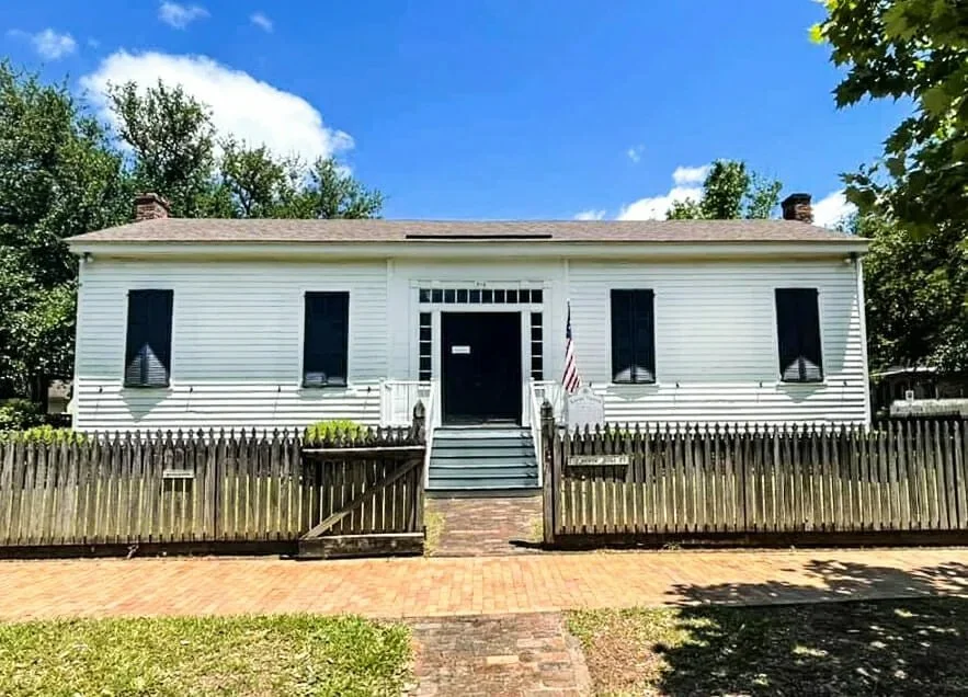A white, two-story house with black shutters, a wooden fence, and steps leading up to the front door, with a small American flag near the entrance, under a bright blue sky with some clouds.