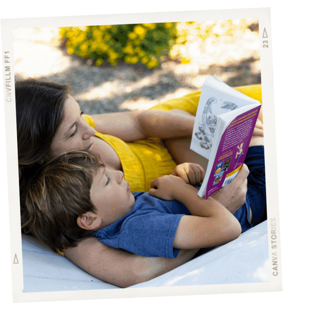 A woman and young boy lie on a blanket outdoors, reading a children's book together, with blurred greenery in the background.