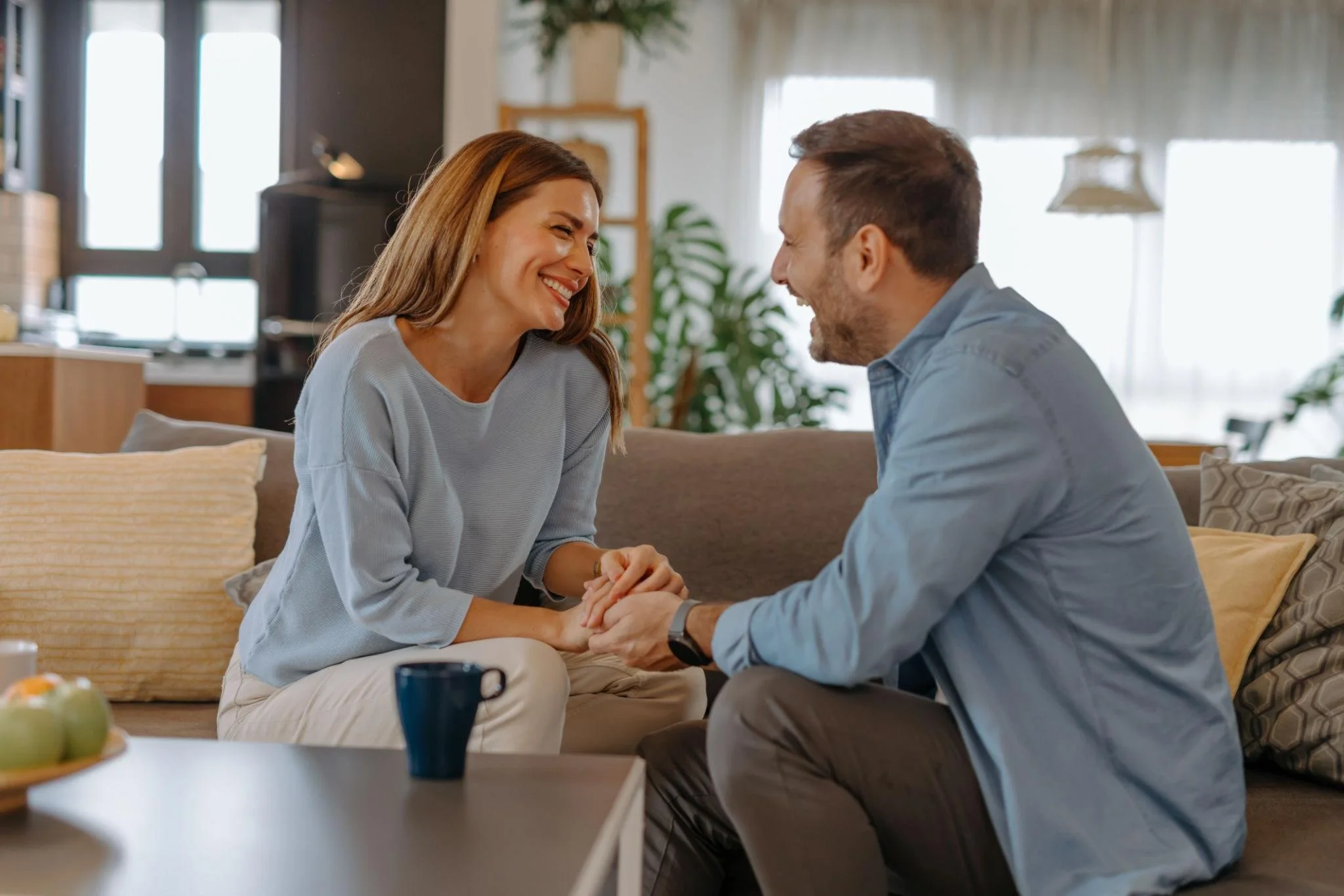 A woman and a man smiling and holding hands, sitting on a couch in a bright living room.