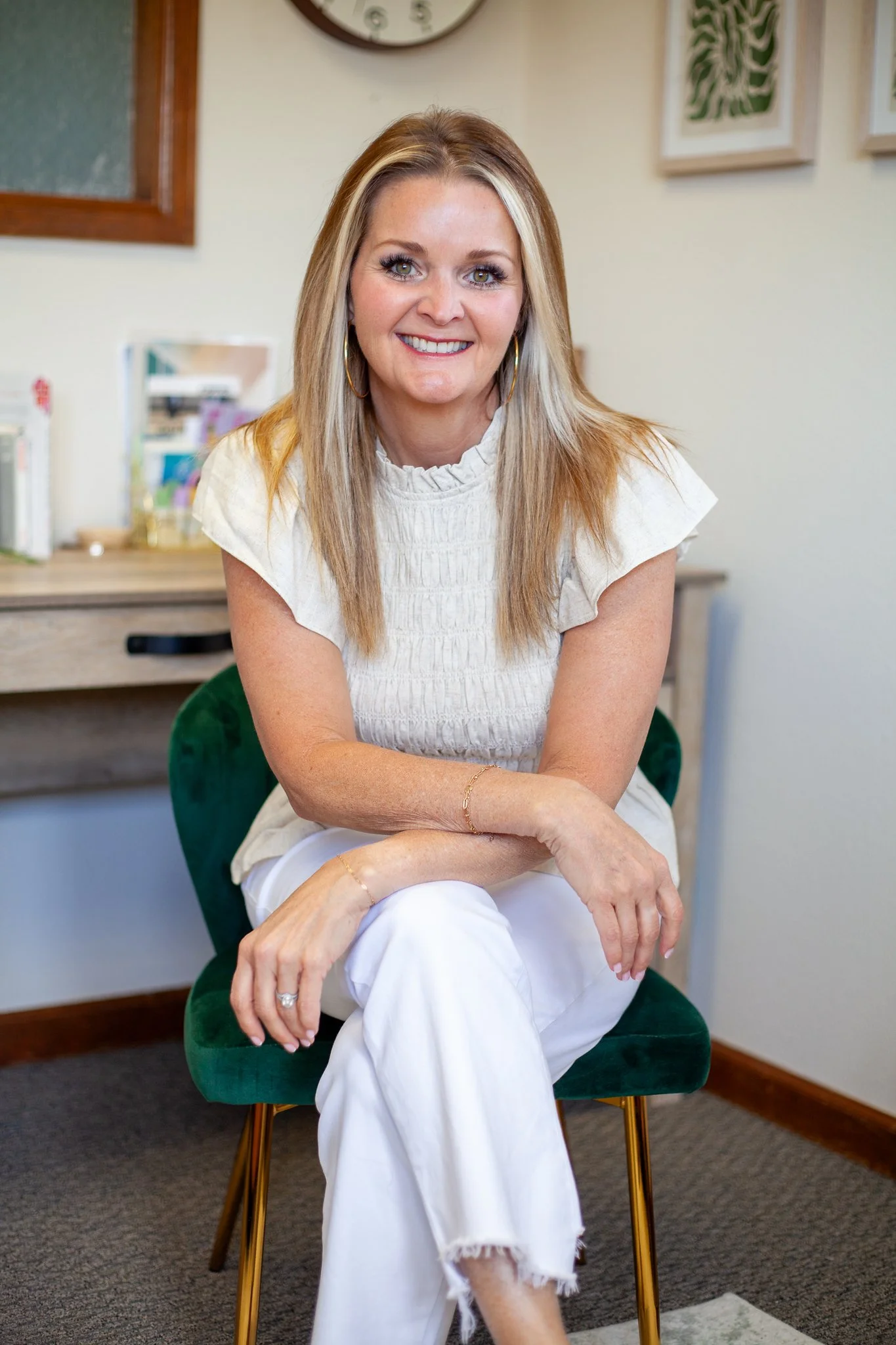 A woman with blonde hair, wearing a white blouse and white pants, sitting in a green velvet chair in an office setting, smiling at the camera. Ready for emotionally focused therapy in colorado springs, CO.