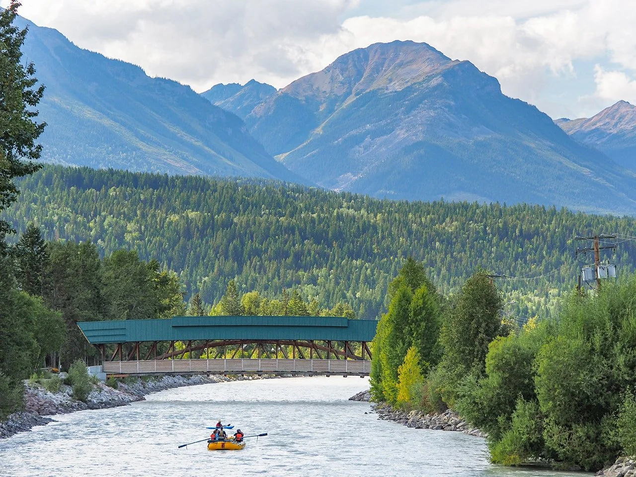 A river flowing through a forest with a mountain range in the background. A bridge crosses the river, and people are kayaking on the water.