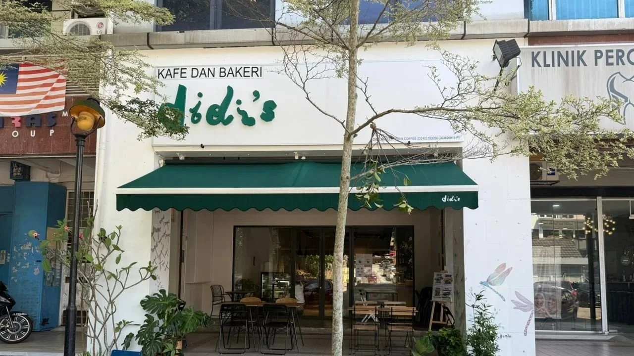 Front view of a bakery and cafe named didi's with outdoor seating, a green awning, and a small tree in front.