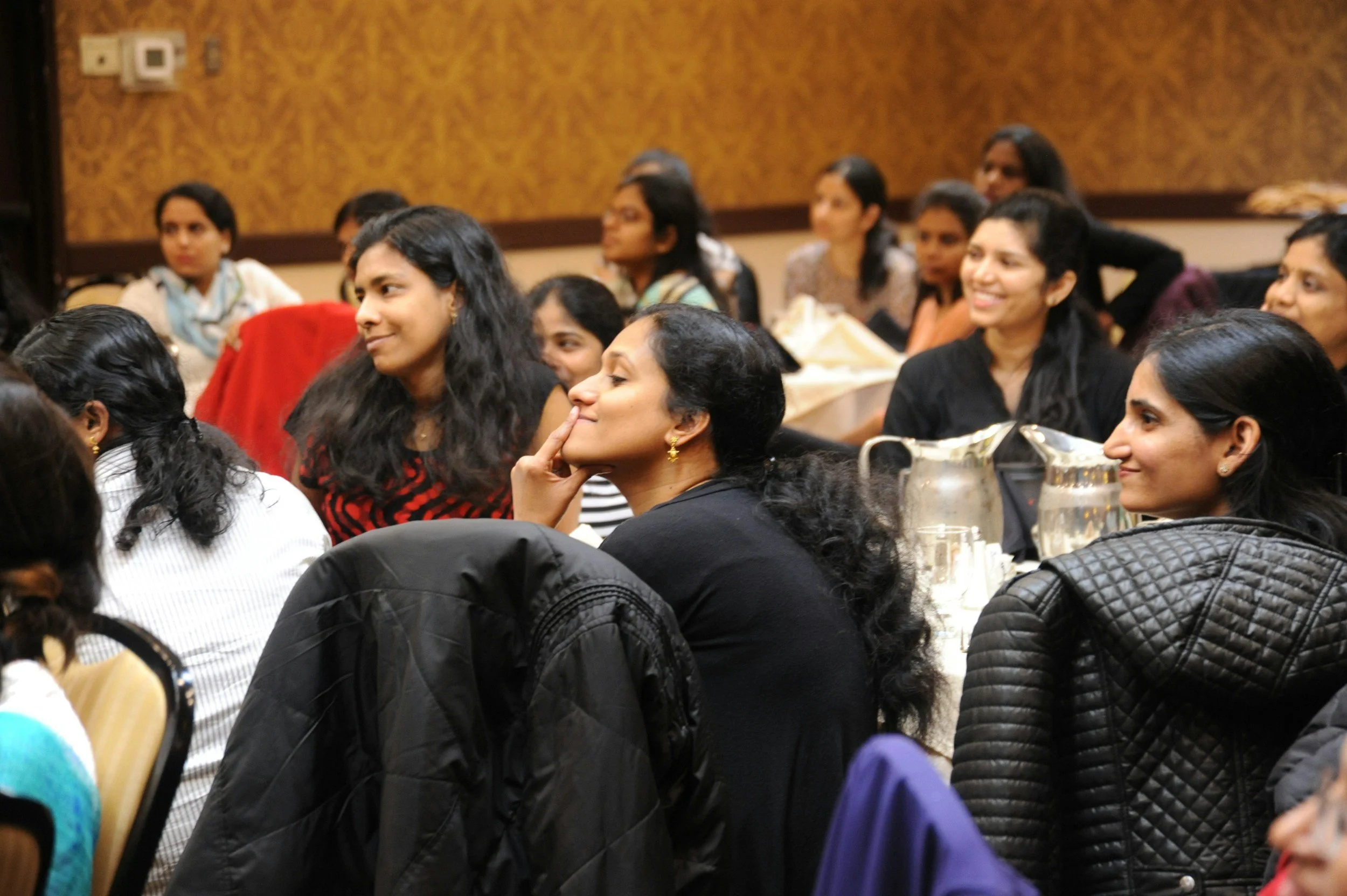 A group of women sitting and listening attentively during a seminar or conference. The room has brown patterned walls and tables with water pitchers.