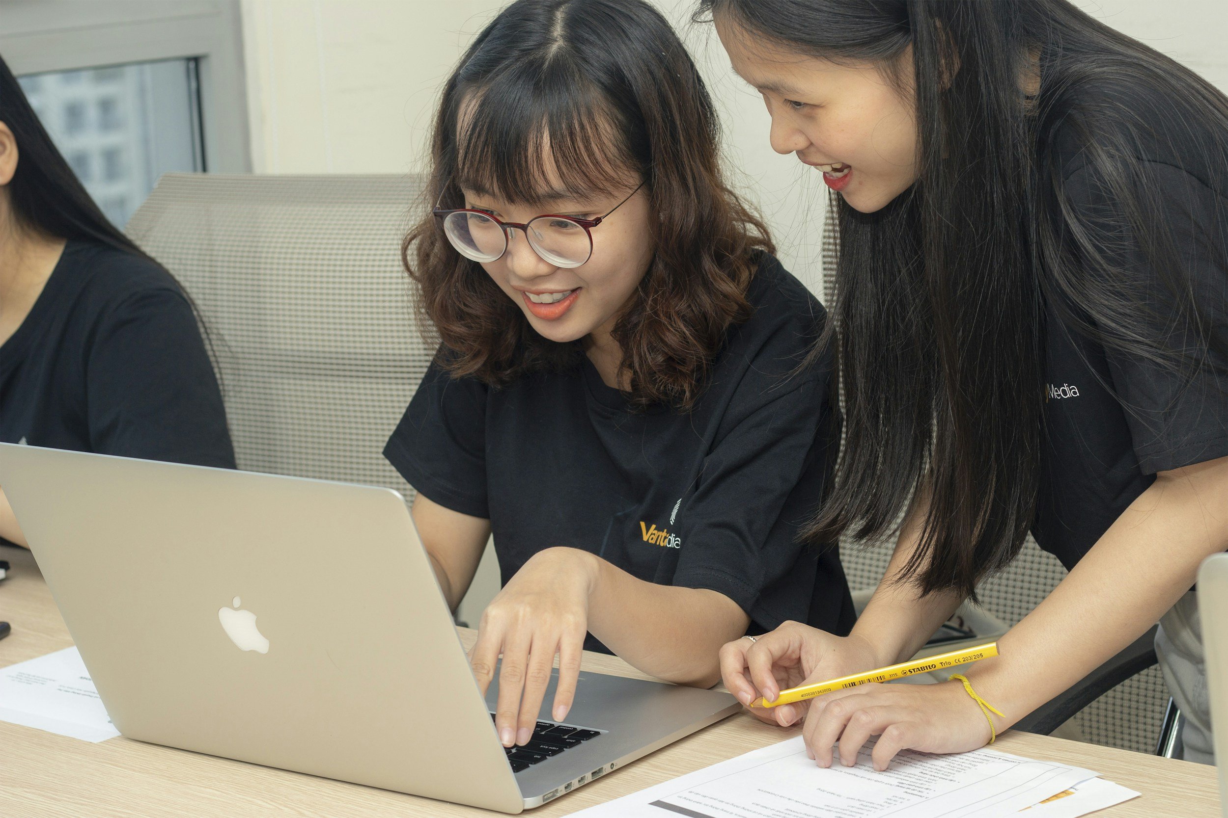 Two women engaging with a MacBook laptop in an office setting, one woman typing and the other pointing at a document with a pencil.