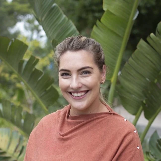 Smiling person in orange top standing in front of large green leaves outdoors