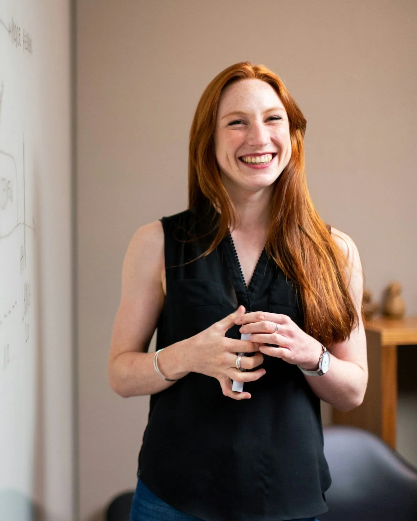 Smiling woman with long red hair wearing a black sleeveless top, standing indoors next to a whiteboard.