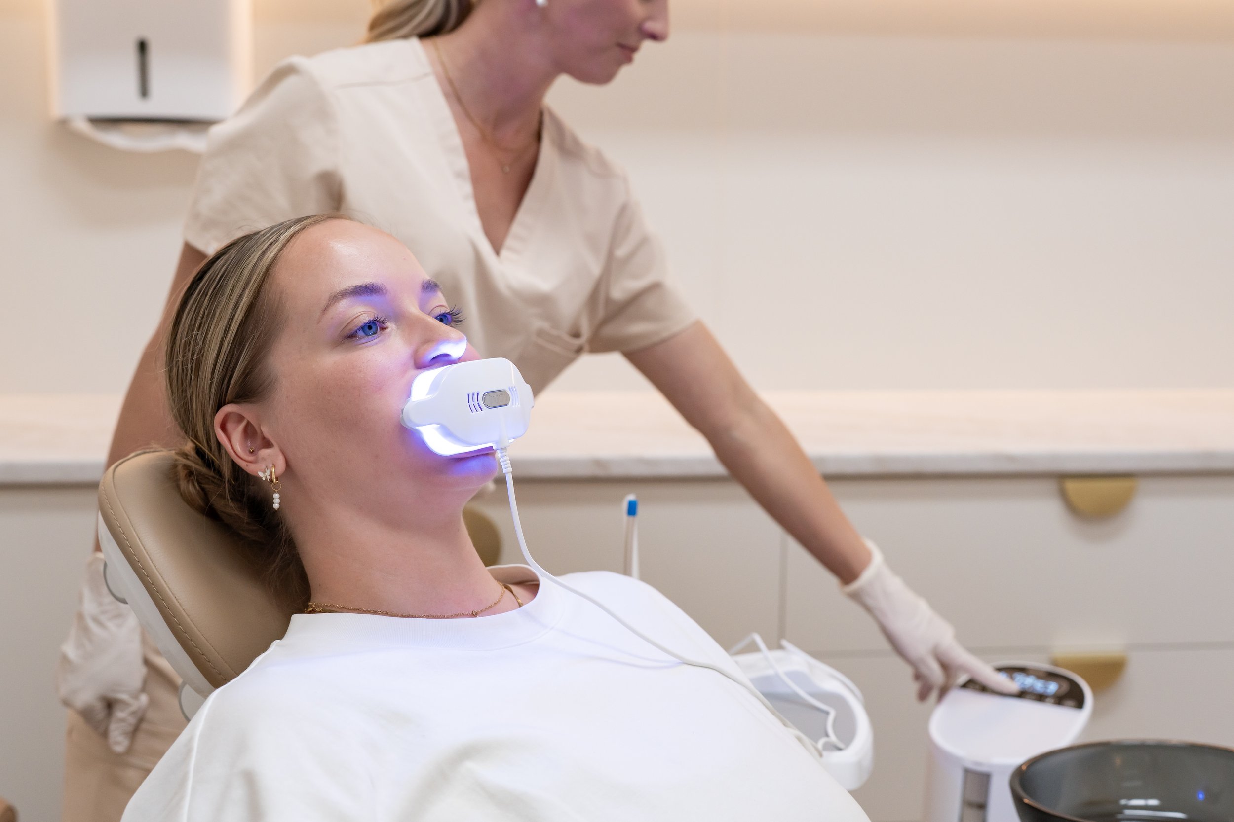 A woman receiving a teeth whitening treatment with a blue LED light on her mouth while sitting in a dental chair, attended by a healthcare professional.