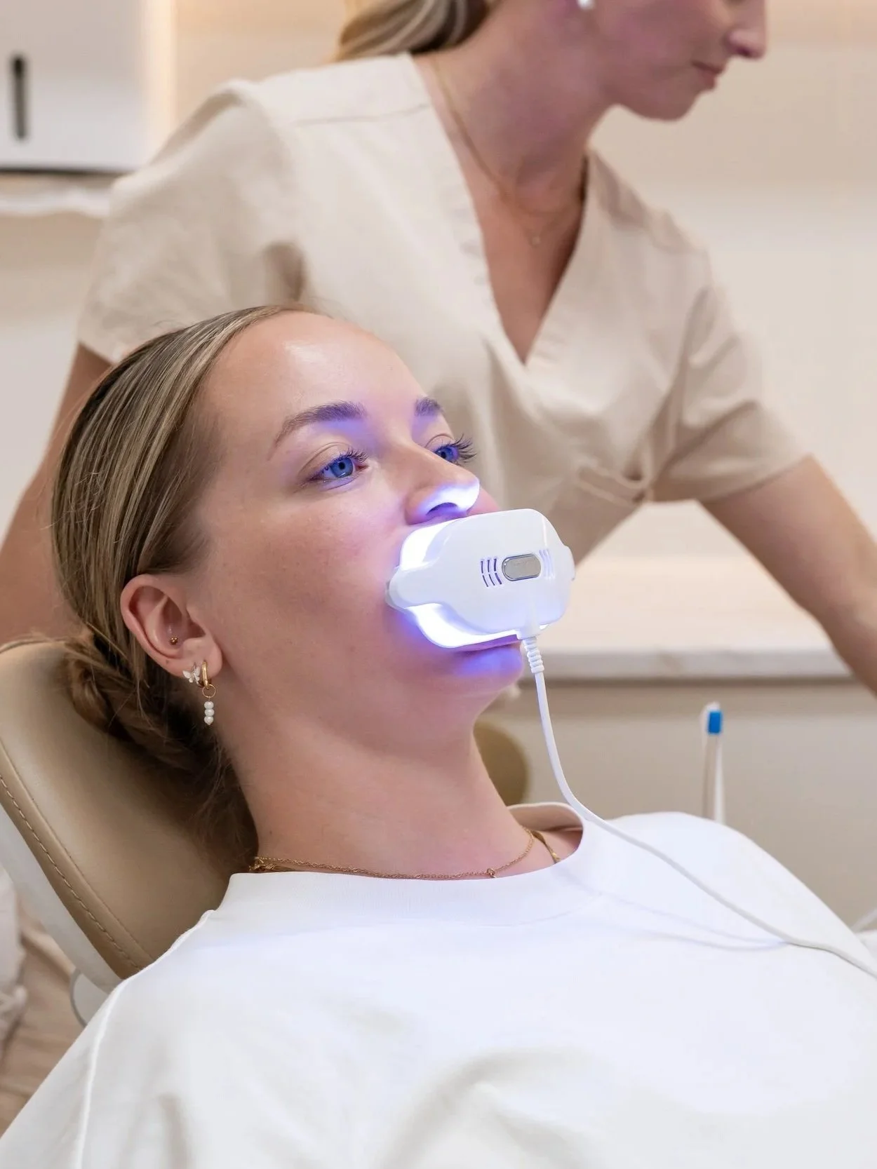 A woman undergoing a cosmetic or dental procedure with a white LED light device under her chin, while a healthcare professional observes in the background.
