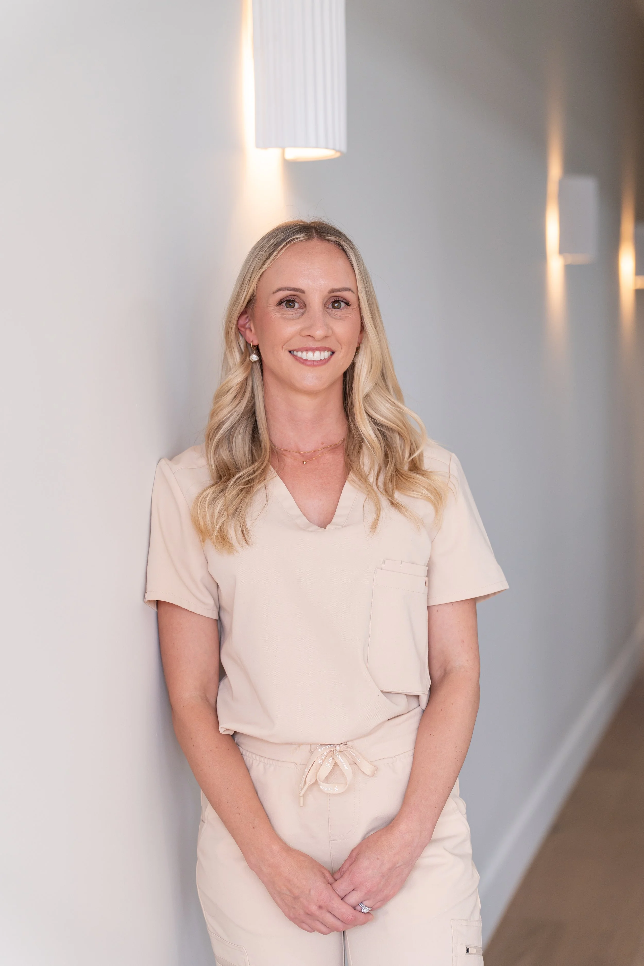 A woman with long blonde hair wearing light-colored scrubs standing against a wall in a well-lit hallway, smiling at the camera.