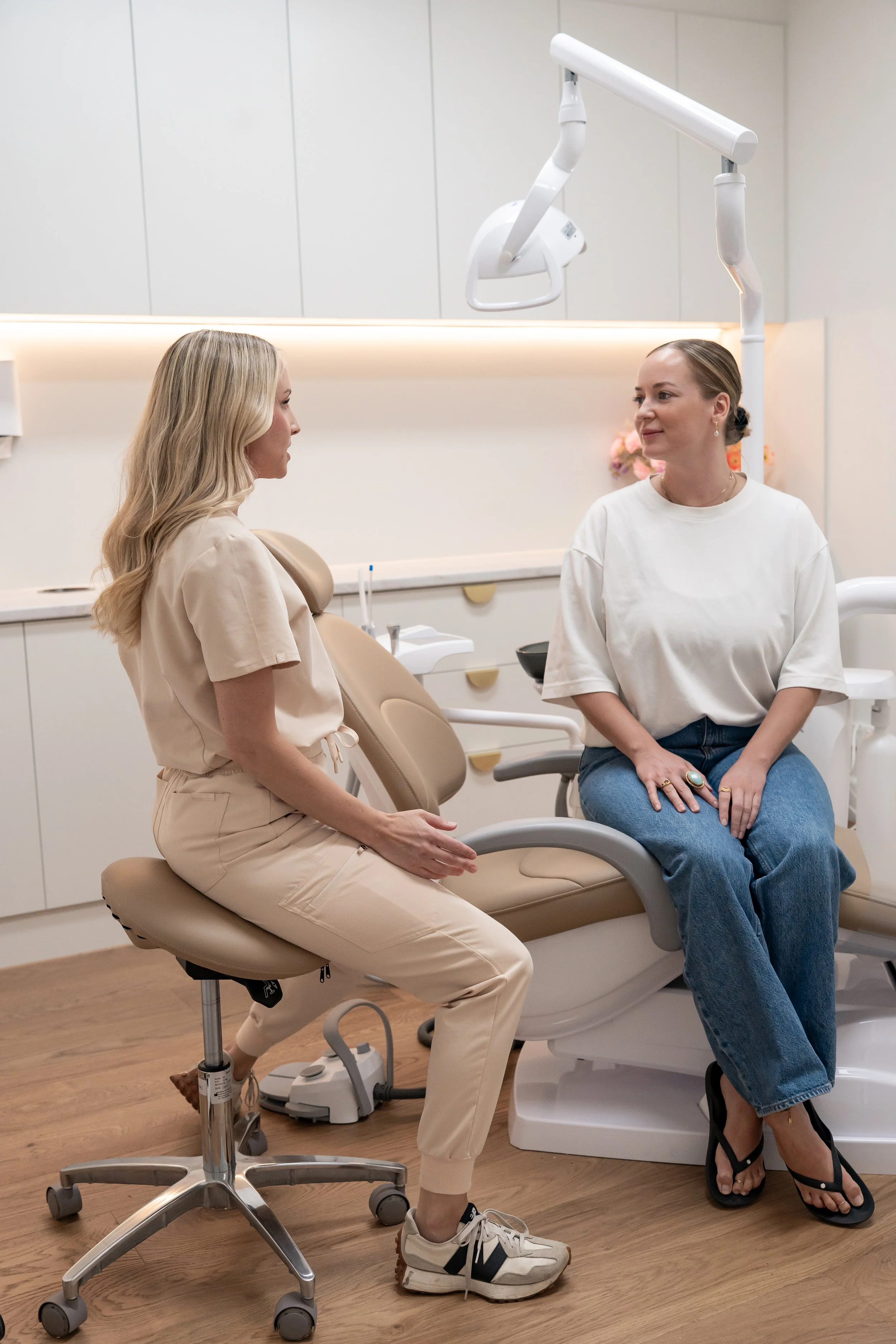 A woman in a white t-shirt and jeans sitting in a dental chair, talking to a dental professional in beige scrubs who is seated nearby, in a modern dental clinic.