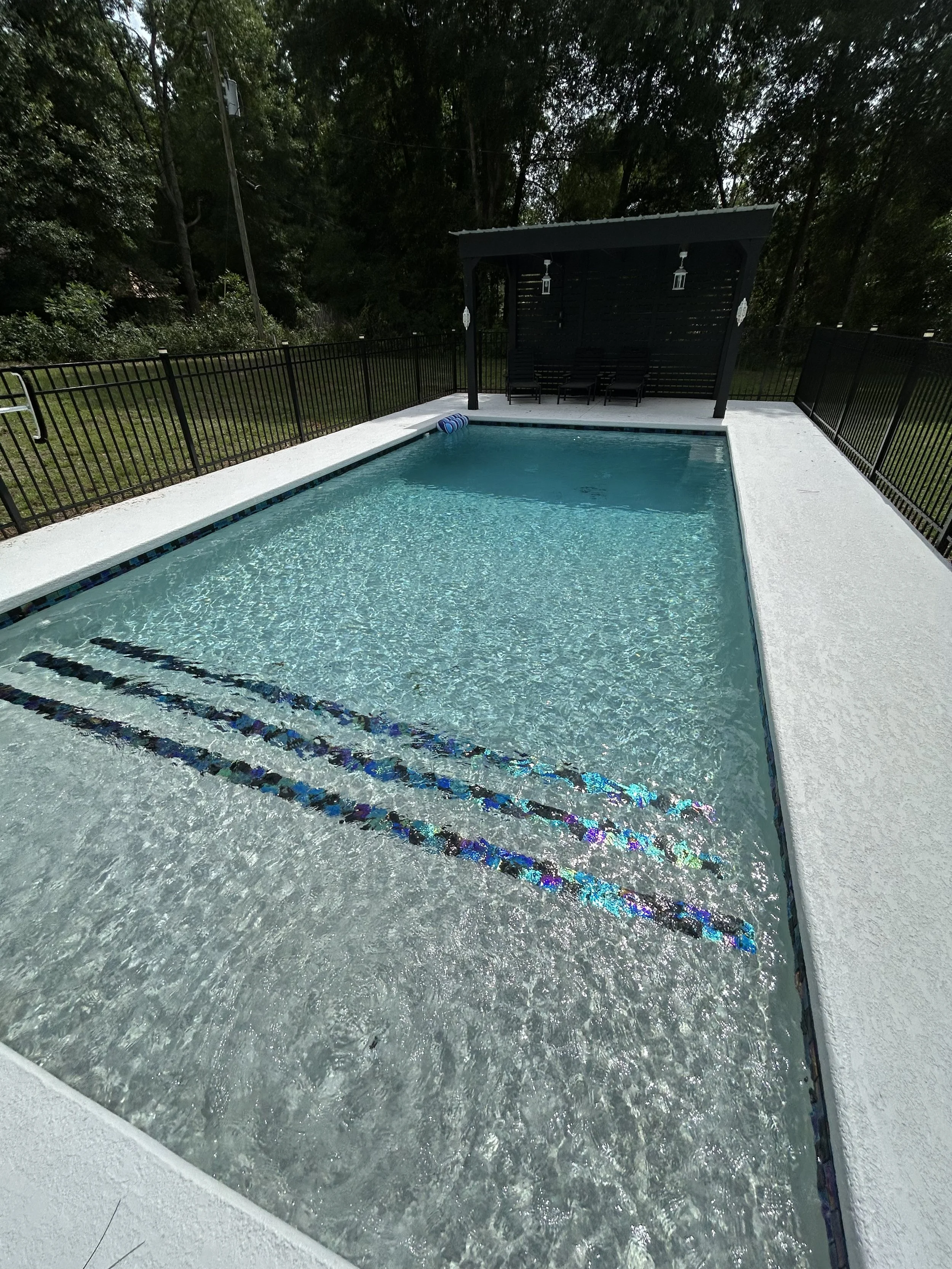 A backyard swimming pool with a black privacy screen and black lounge chairs, surrounded by a black metal fence, with trees and a power pole in the background.