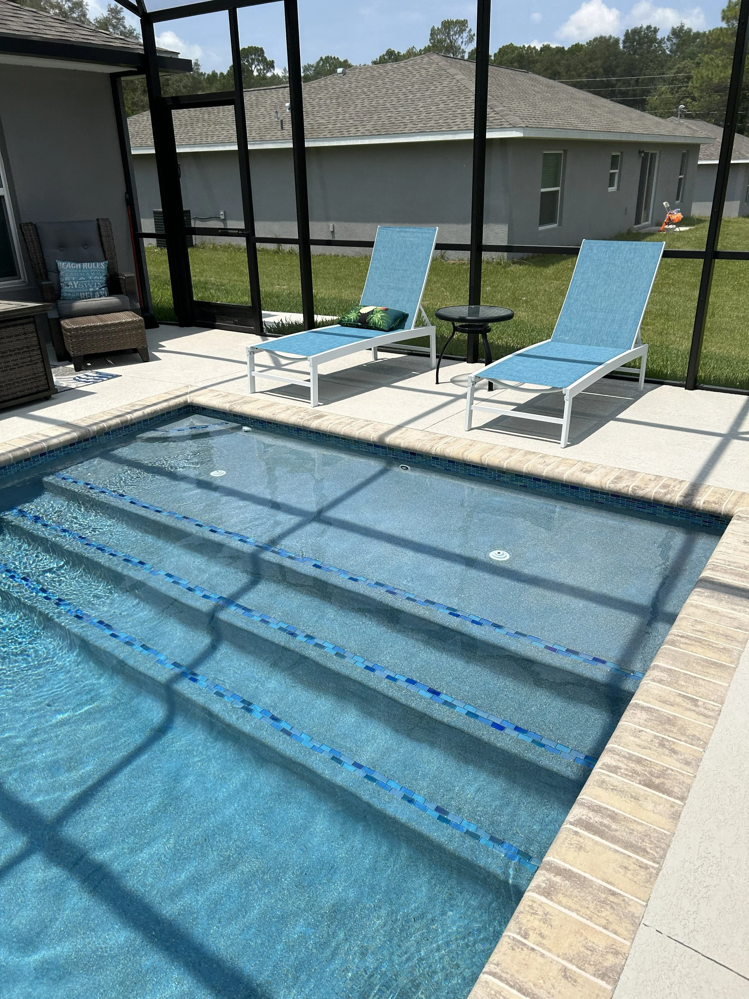 View of a backyard swimming pool enclosed by a screened patio with blue lounge chairs, a small black table, and a wicker chair. In the background, there is a lawn and house with a gray roof.