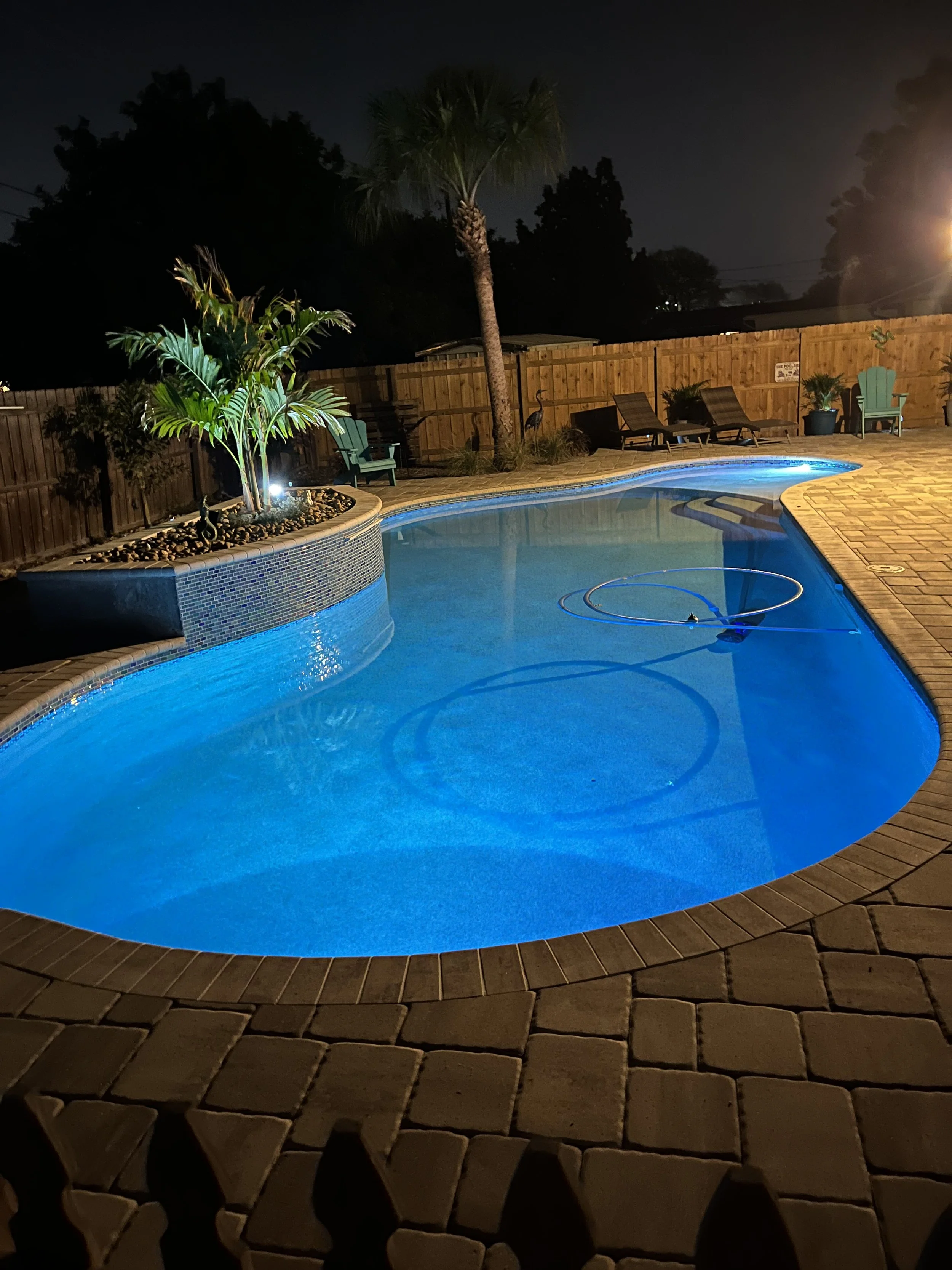 Dark backyard at night featuring a lit swimming pool with blue lighting, surrounded by a brick patio, lounge chairs, potted plants, and a tall palm tree. A wooden fence encloses the area.