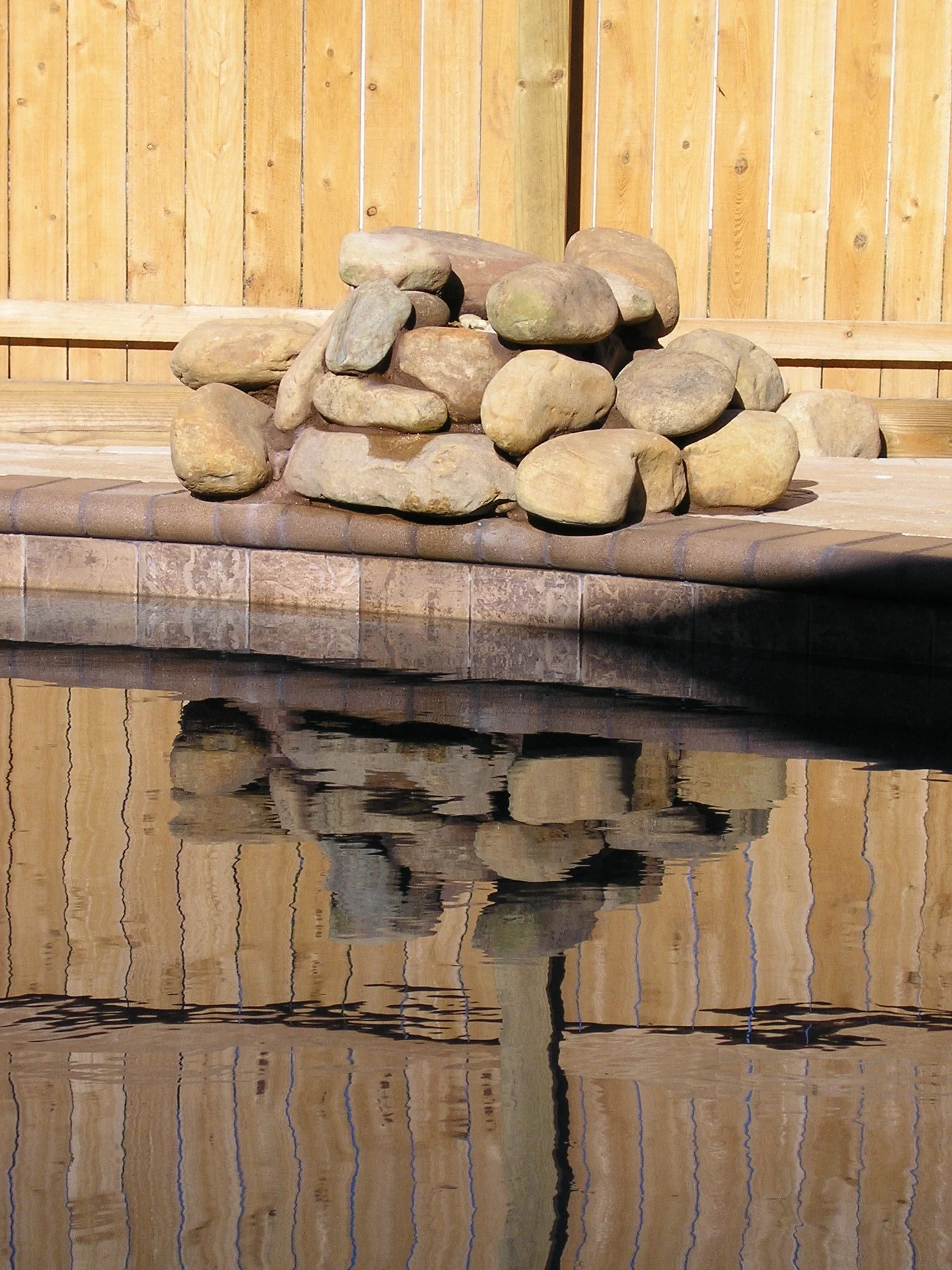 A rock formation on a patio with a wooden fence in the background, and its reflection in a body of water.