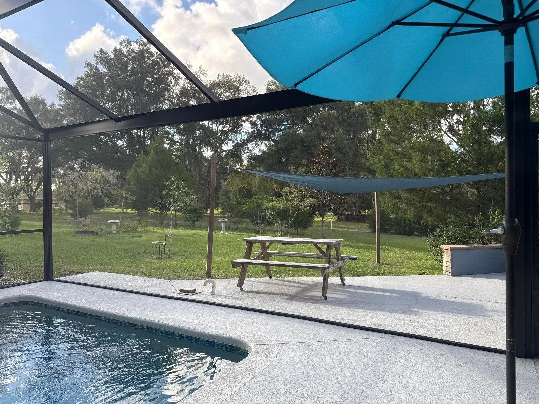 View of a backyard swimming pool area with a screened enclosure, a picnic table with a bench, a blue umbrella shading a lounge chair, and a grassy yard with trees and birdbaths in the background.