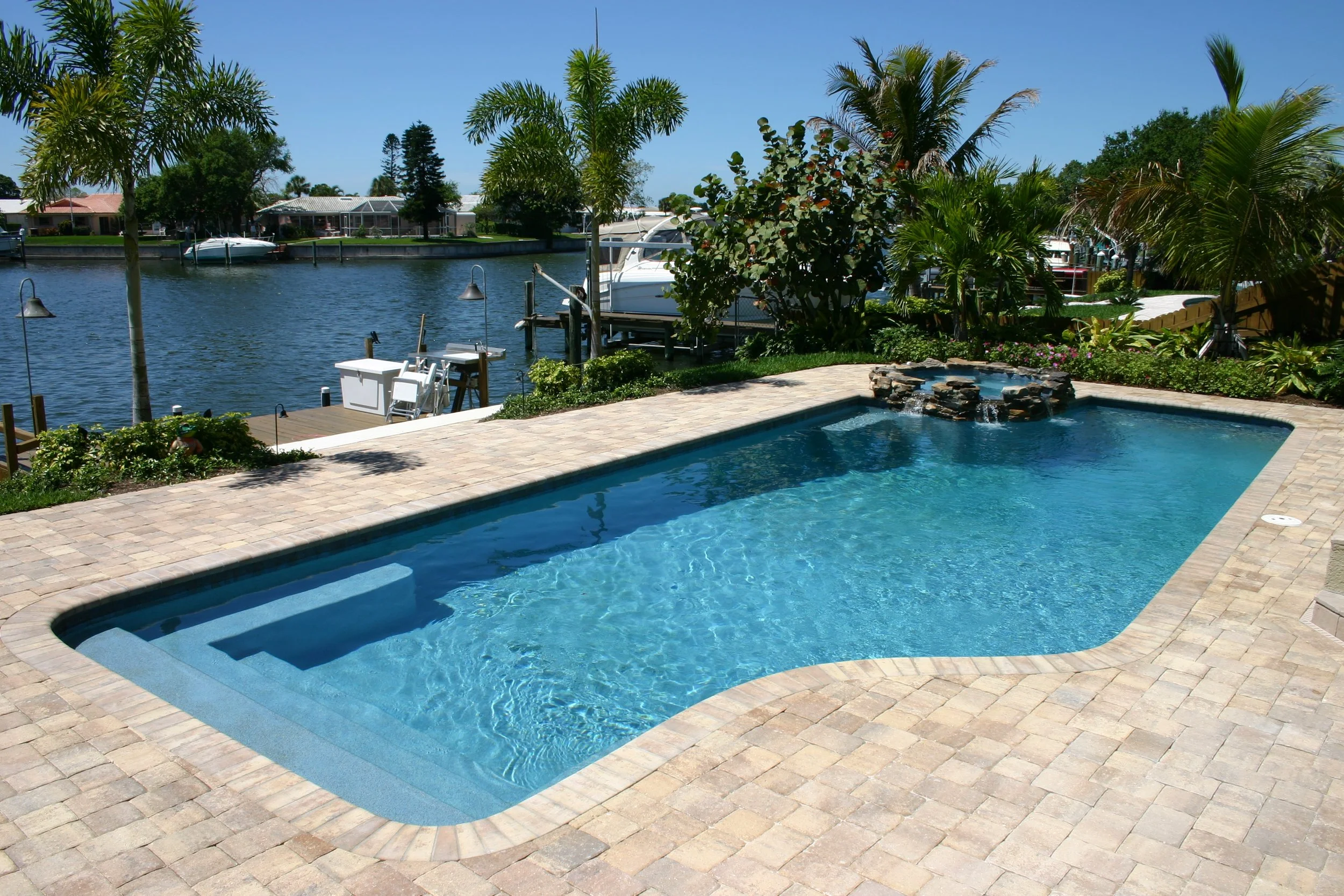 Backyard pool with waterfall feature, surrounded by stone pavers, overlooking a canal with boats and houses with lush greenery and palm trees.