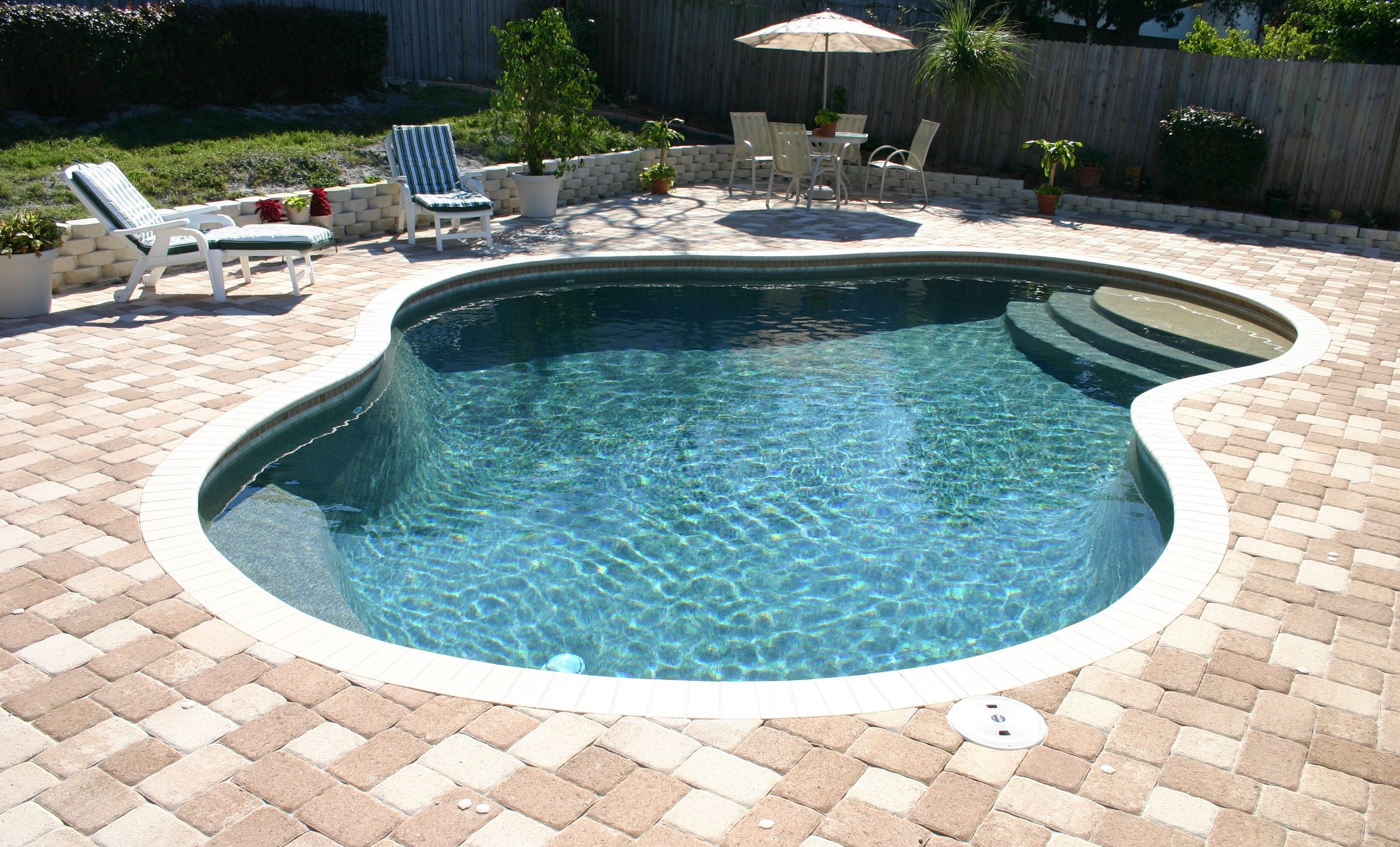 Residential backyard with an in-ground pool, brick patio, outdoor seating area with chairs and table under a patio umbrella, potted plants, and a wooden fence.