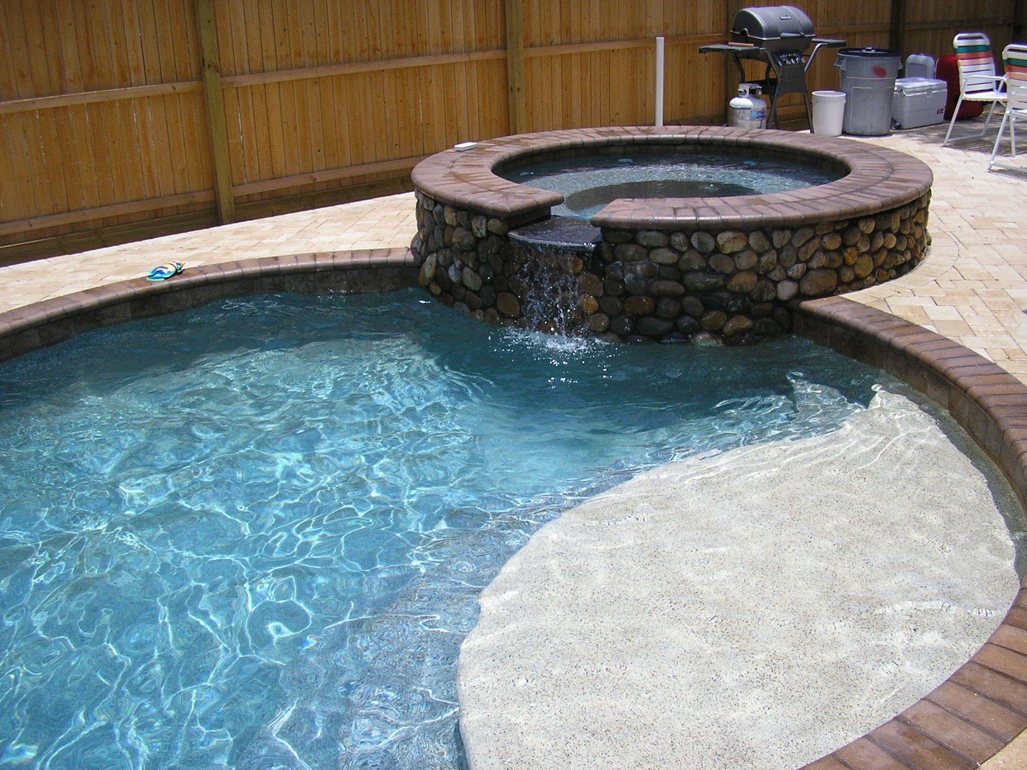 Above ground hot tub with stone exterior on a paved patio, adjacent to a swimming pool, enclosed by a wooden privacy fence, with patio furniture and grilling equipment in the background.