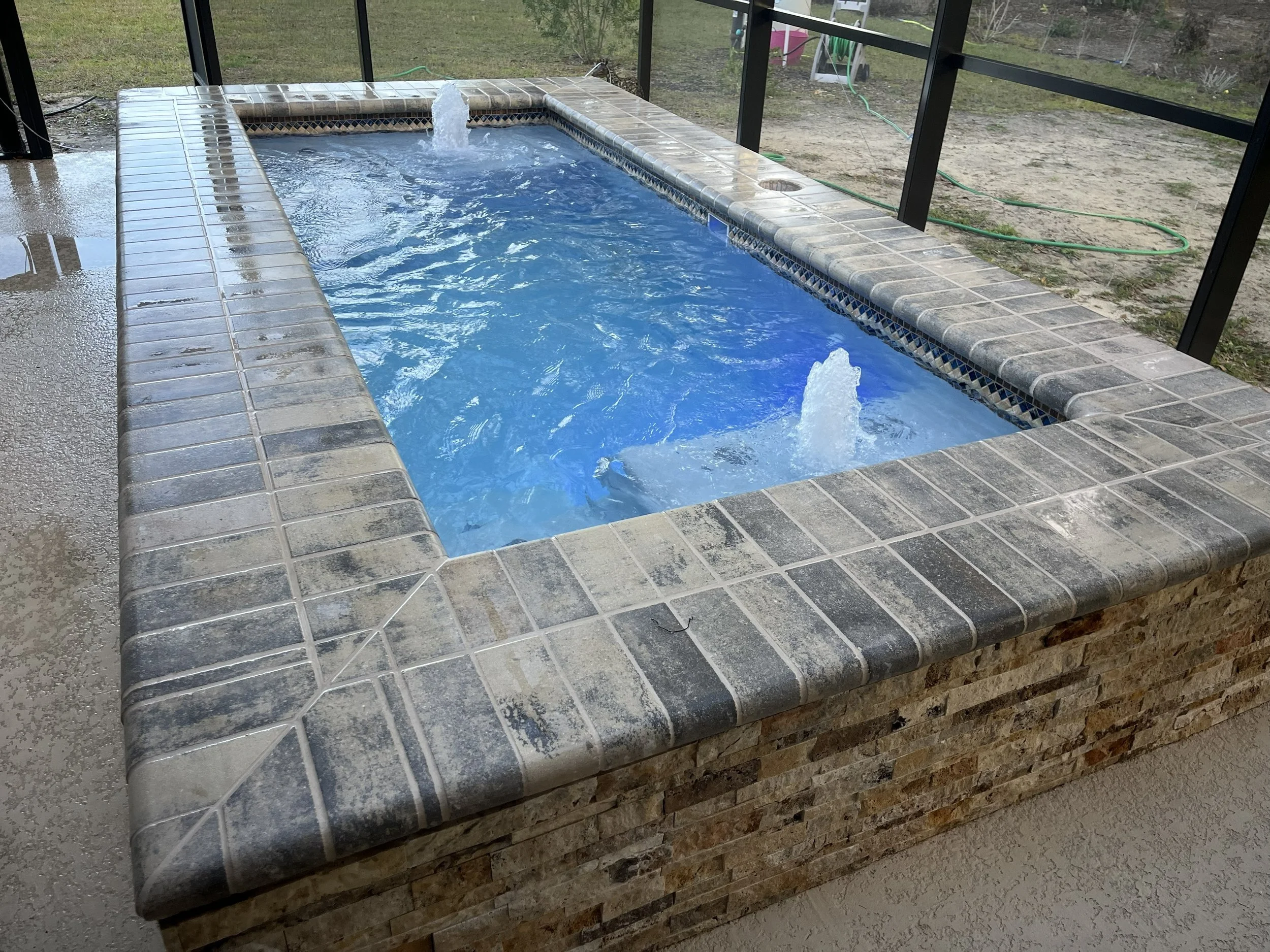A rectangular hot tub with water jets and a small waterfall feature, surrounded by a tiled border, located on a patio with a screen enclosure.