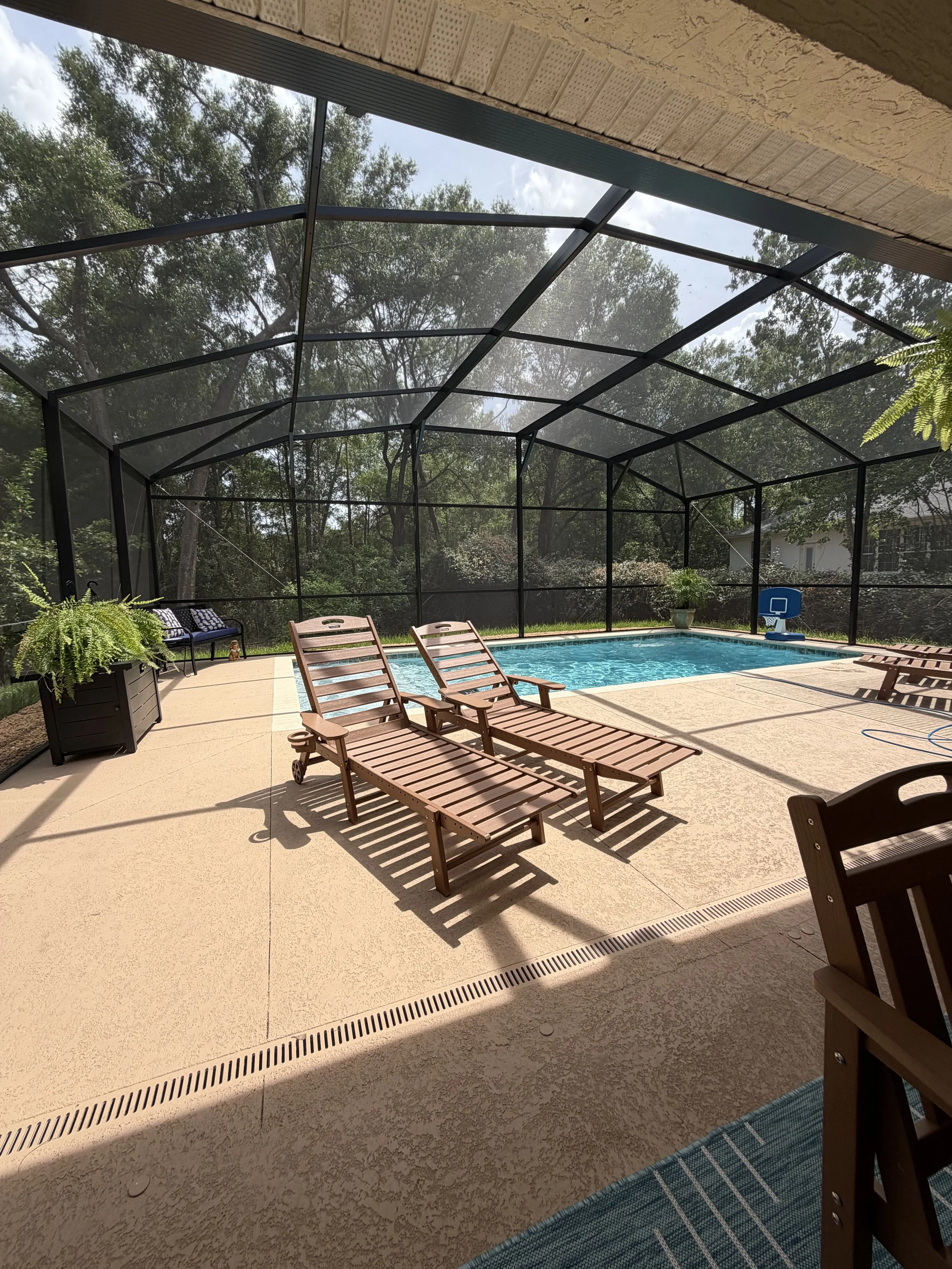 Patio area with lounge chairs, potted plants, and a swimming pool enclosed by a screened structure, with trees in the background and a part of the house visible.