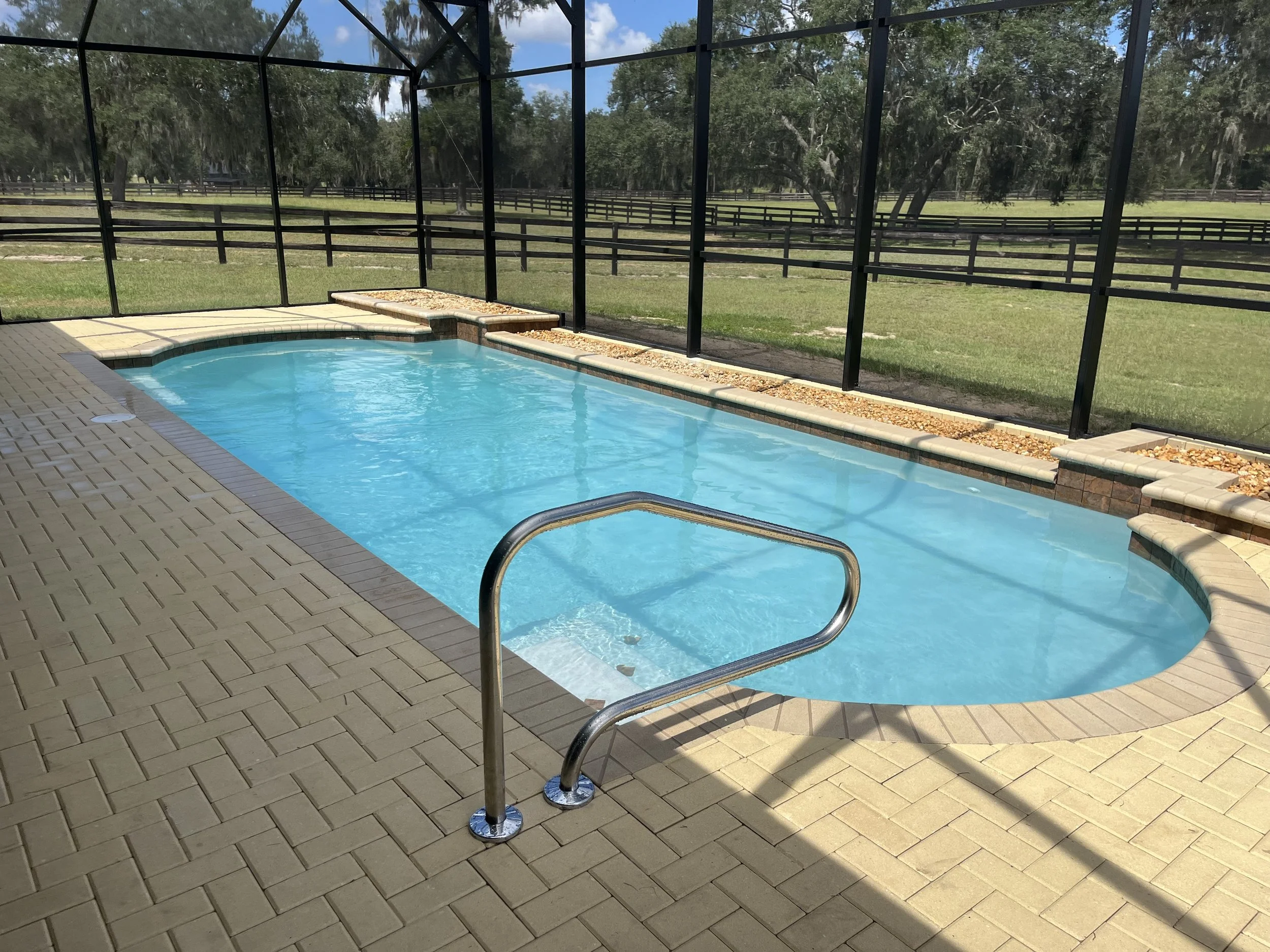A small, curved swimming pool with blue water, surrounded by brick and beige tile decking. The pool is enclosed by a black metal and screen enclosure, overlooking a grassy area with trees and fencing beyond.
