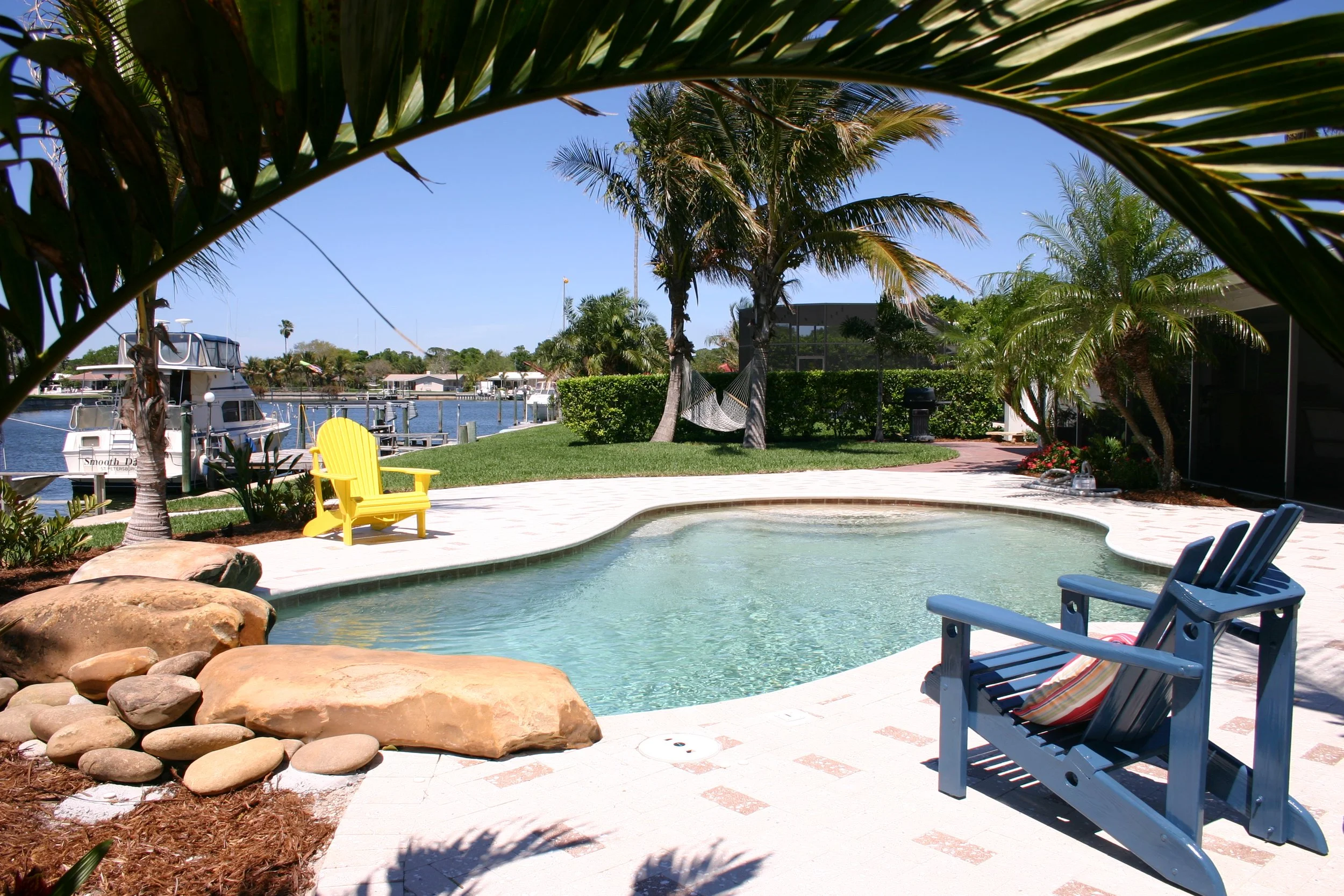 A backyard with a small pool, blue chairs, a yellow chair, palm trees, a hammock, and boats docked at a marina in the background under a clear blue sky.