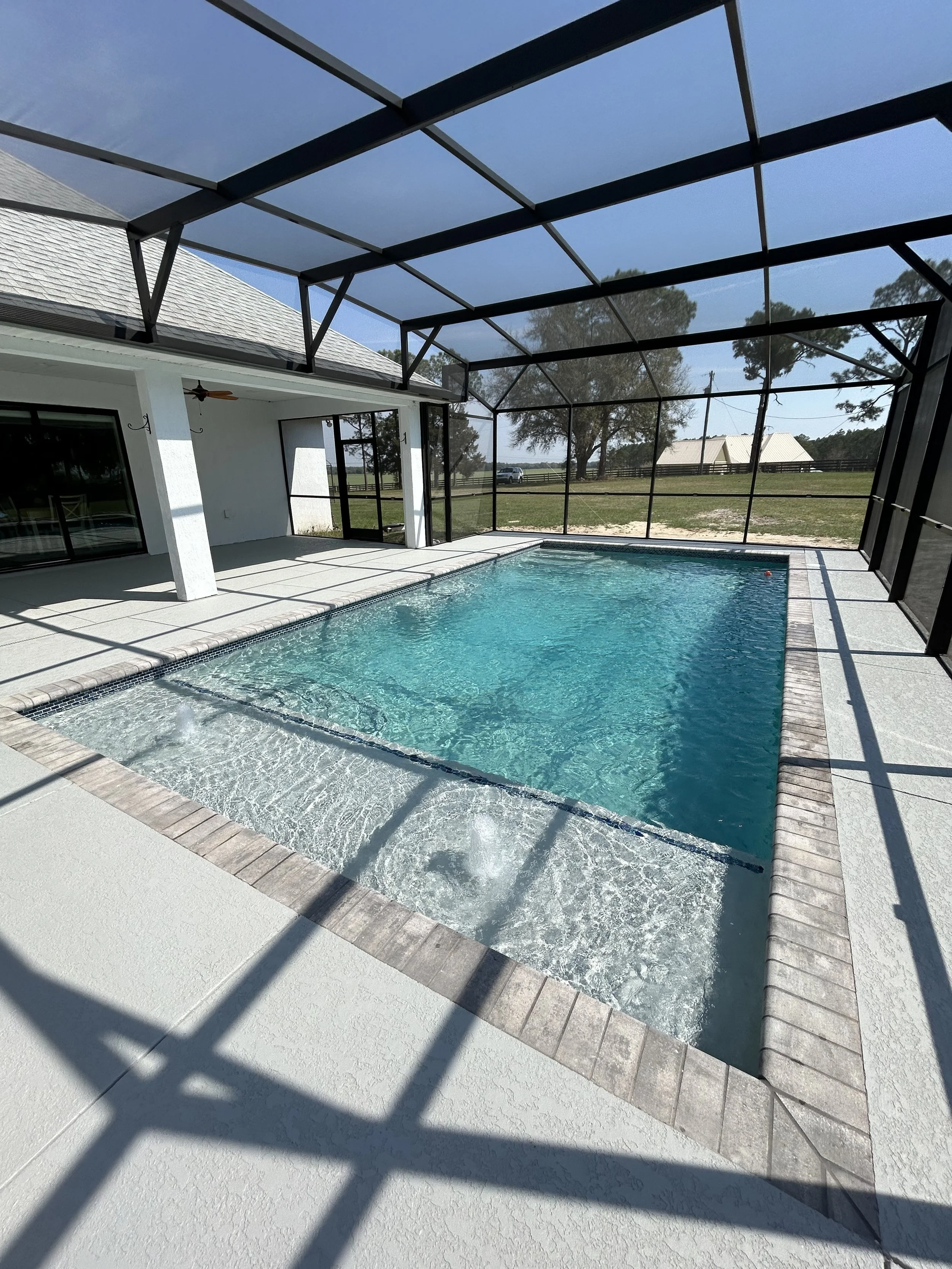 A backyard swimming pool enclosed by a screened structure with black framing, next to a white house with sliding glass doors and windows, and a backyard with lawn, trees, and a white barn in the distance.