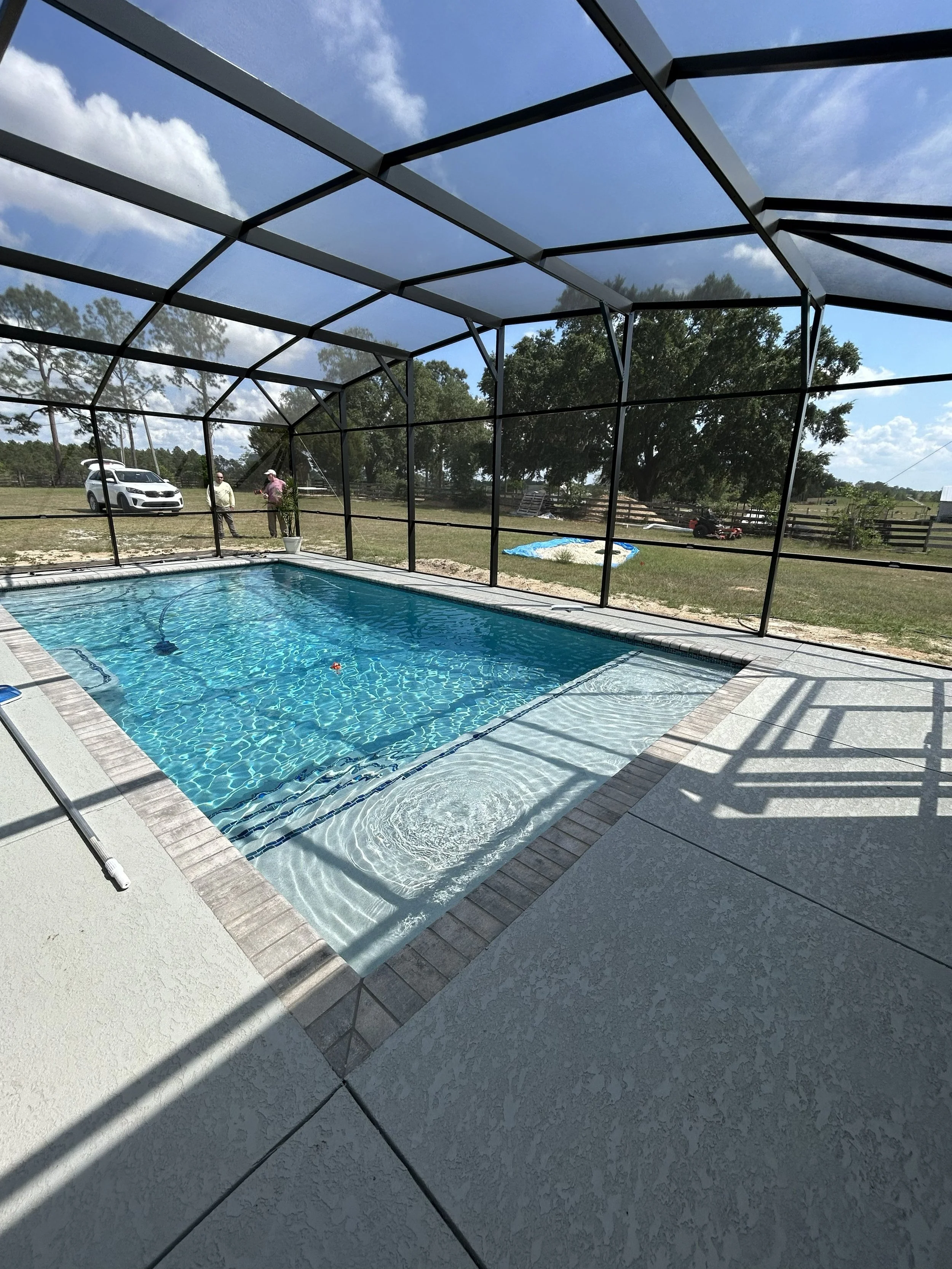 View of a backyard swimming pool enclosed by a screen structure with a blue sky and scattered clouds overhead. Two people are talking near the pool, and a white car is parked in the background.