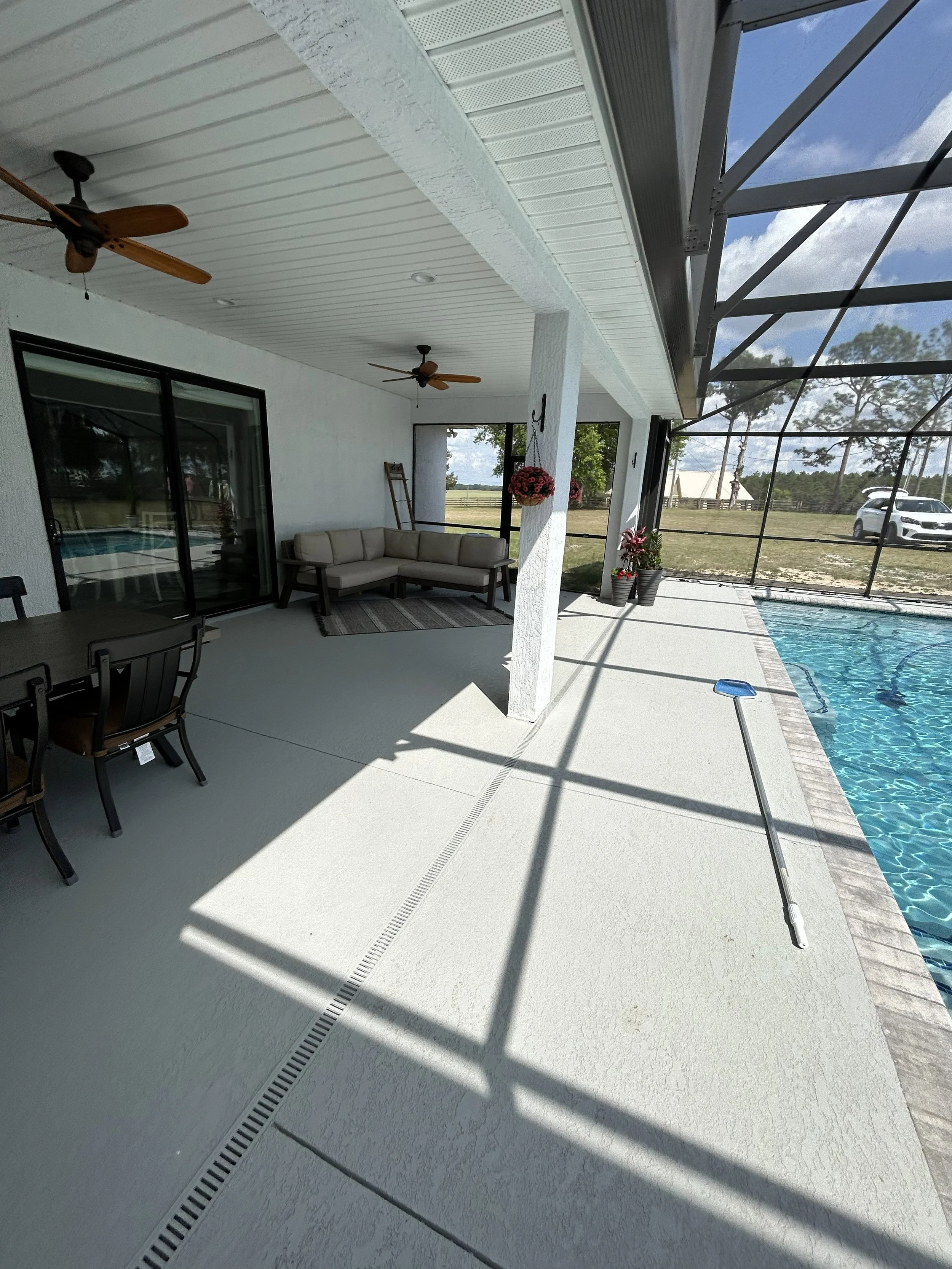 View of a screened-in backyard pool area with white textured concrete floor, a white ceiling with two ceiling fans, a sliding glass door, potted plants, hanging flower baskets, and a clear blue sky with clouds visible through the screen enclosure.