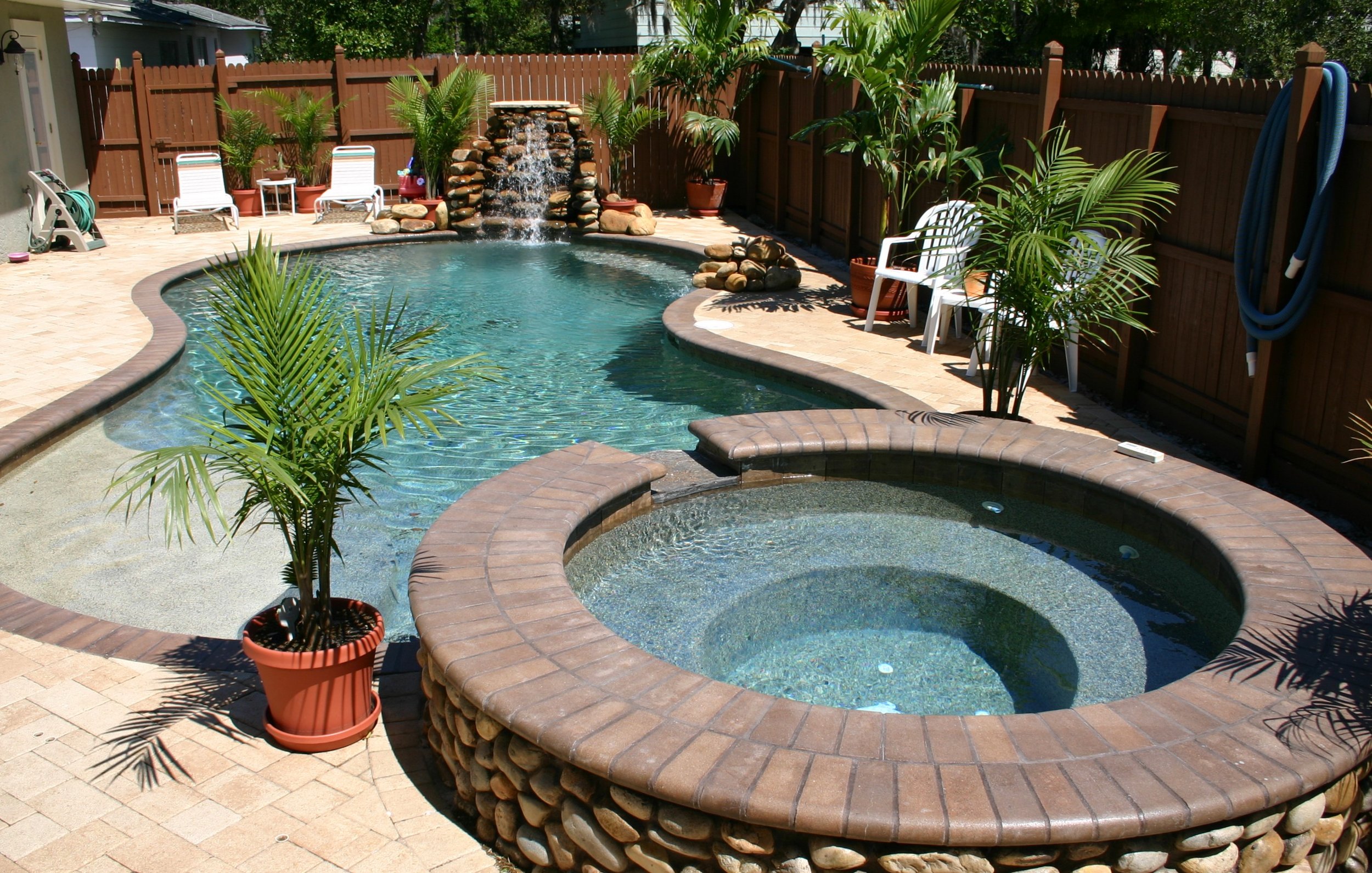 Backyard with swimming pool, hot tub, potted palm plants, and a wooden privacy fence. There are patio chairs, a small table, a garden hose, and a waterfall feature with rocks.