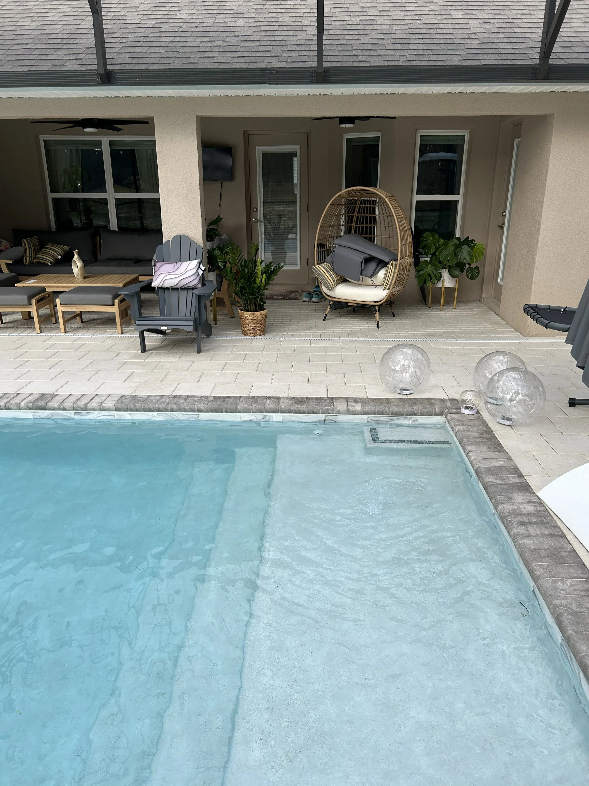 A backyard with a swimming pool, outdoor seating area with gray and wooden furniture, potted plants, a hanging rattan chair with cushions, and a wall-mounted TV, with a screened porch above.