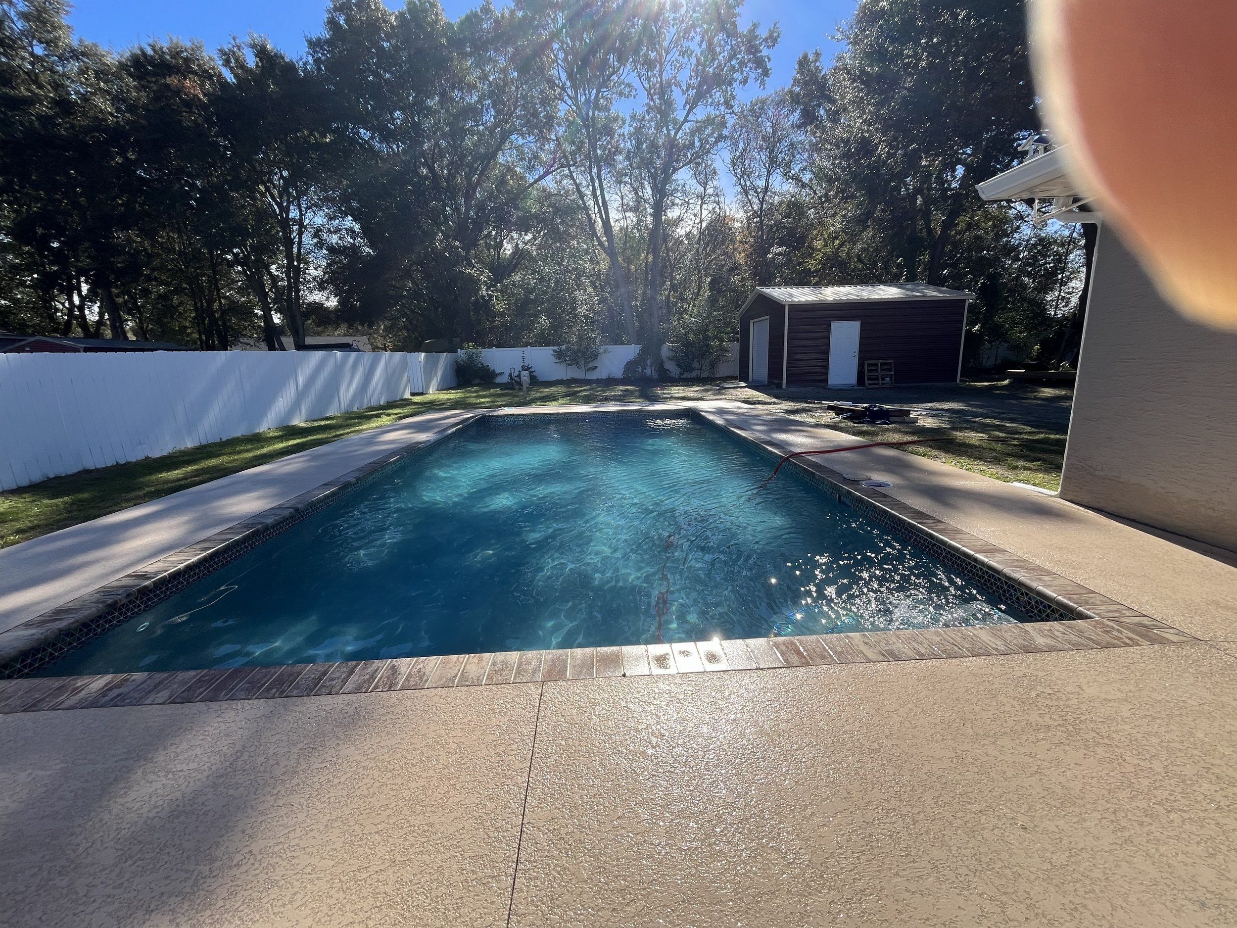 Backyard with a swimming pool, white fence, trees, a small shed, and a house on the right