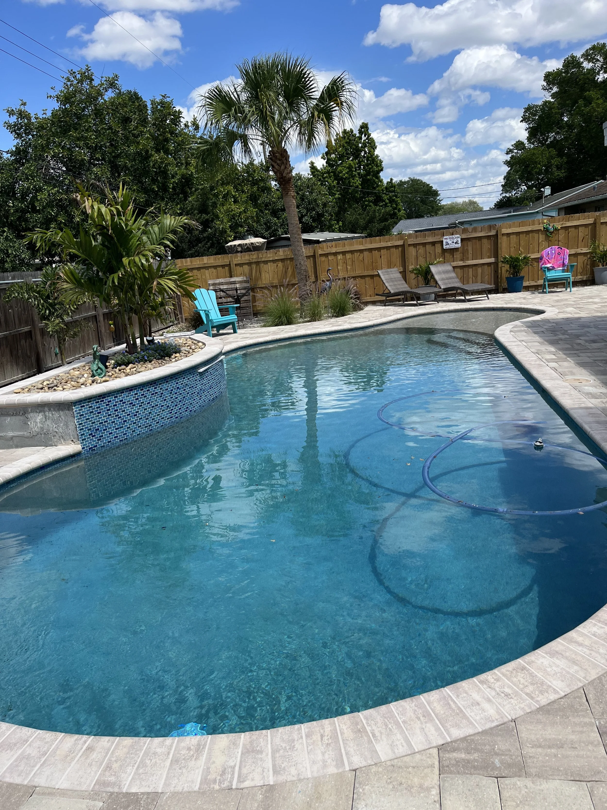 Residential backyard with a swimming pool, lounge chairs, potted plants, a wooden fence, and a palm tree under a partly cloudy sky.