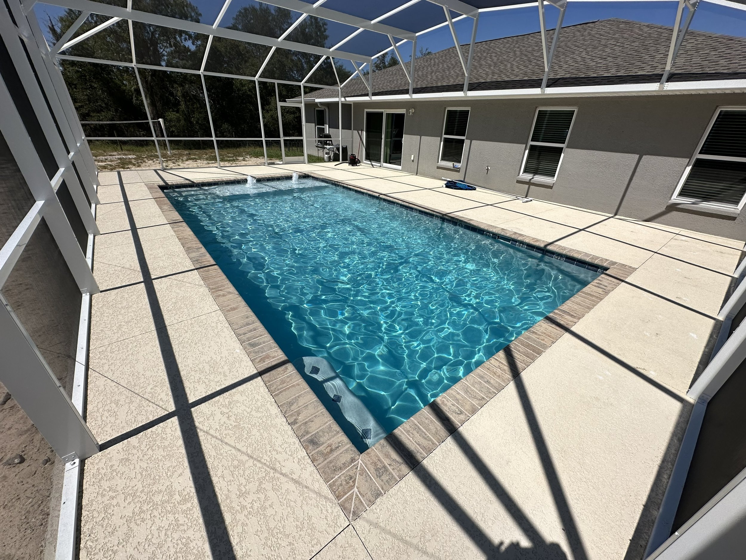 A backyard swimming pool enclosed by a white metal and glass screened patio, with a house wall featuring windows and a sliding glass door in the background, under a sunny sky.