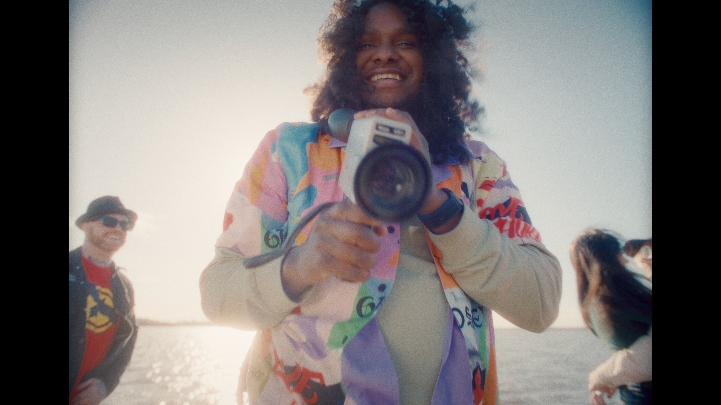 Indigenous artist Baker Boy holds a super 8 film camera in his music video My Mind. He is standing outdoors near a body of water during daylight, with two people in the background.