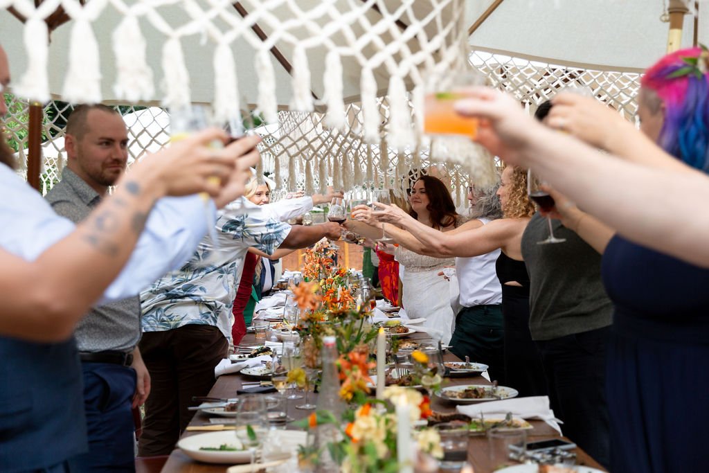 People raising drinks in a toast around a long table decorated with flowers at an outdoor celebration.