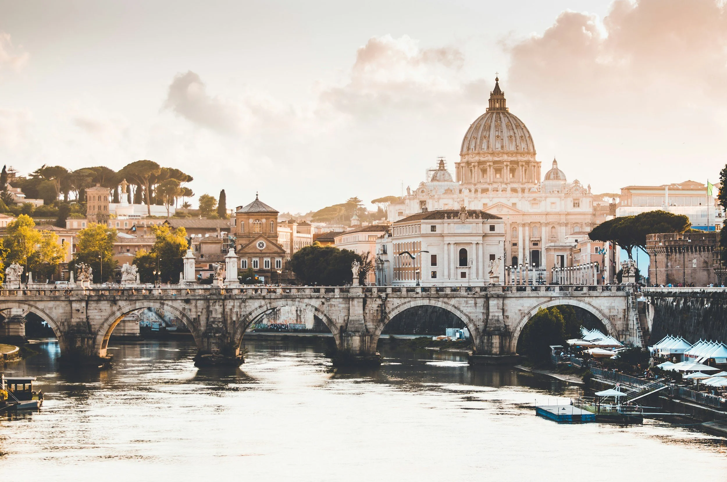 View of St. Peter's Basilica and the Tiber River in Rome, Italy, with historic buildings, a stone bridge, and boats in the foreground during sunrise or sunset.