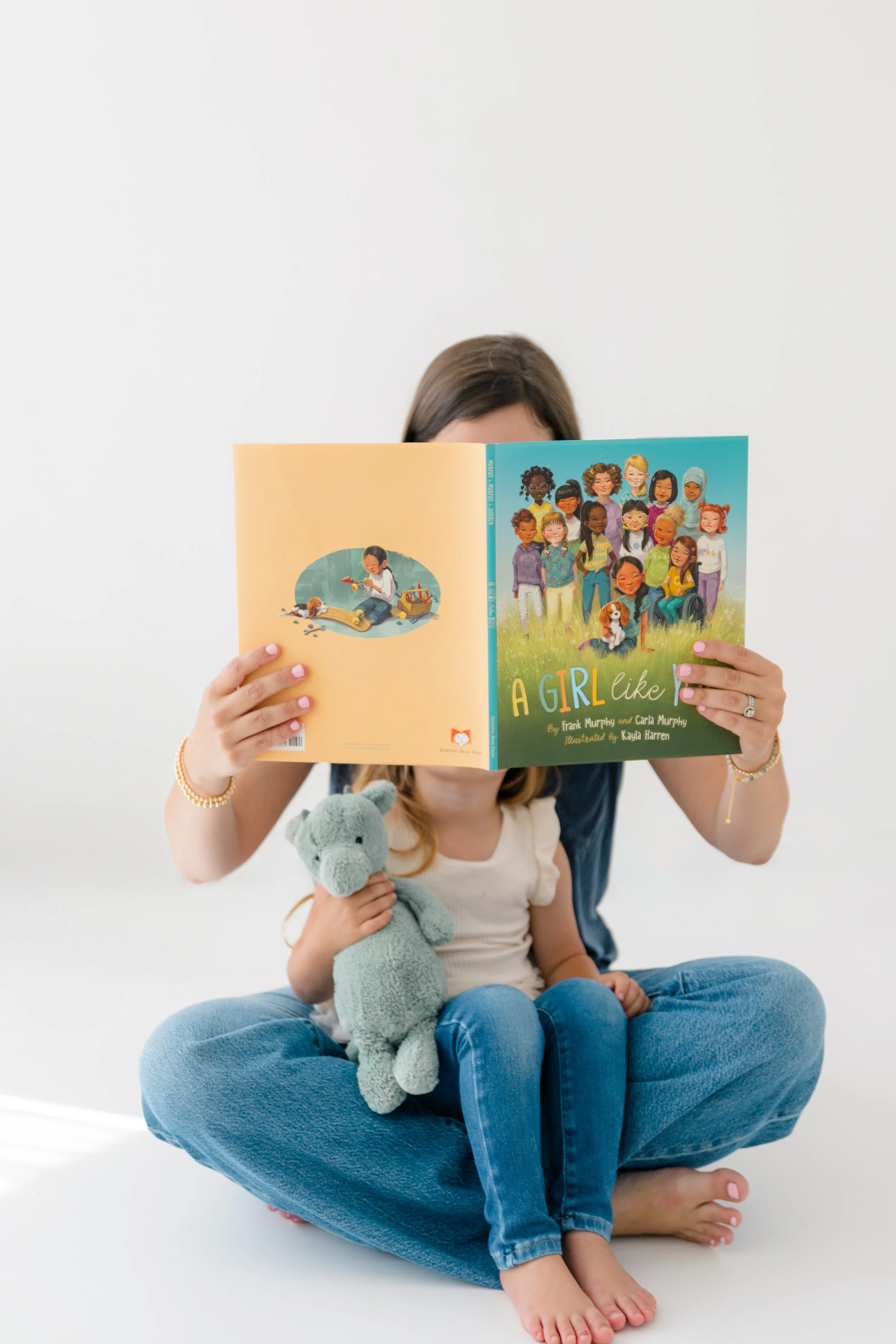 A woman sitting cross-legged on the floor reading a children's book called "A Girl Like" to a young girl, who is holding a plush toy of a gray hippopotamus.