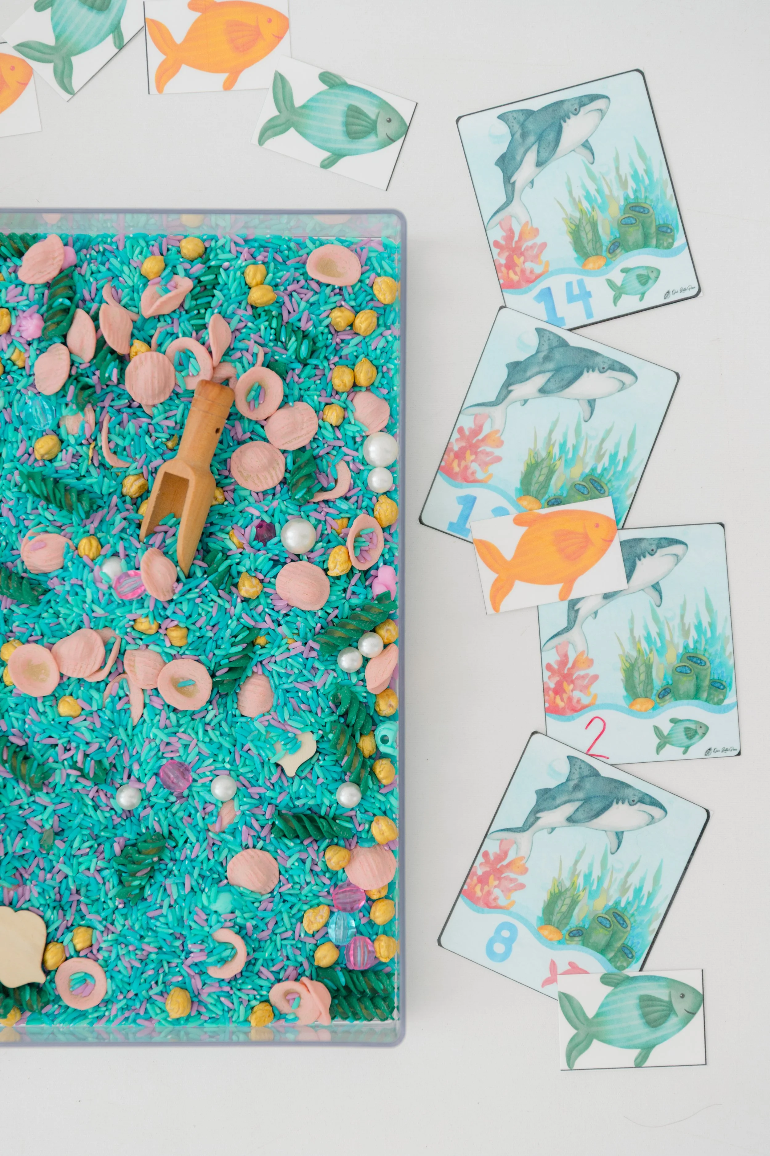 Colorful sensory bin filled with pink, yellow, and purple rice and various small plastic sea animal figures, with a small wooden scoop and decorative pearls scattered inside, alongside fish-themed playing cards featuring illustrations of sharks, fish, and underwater scenes.