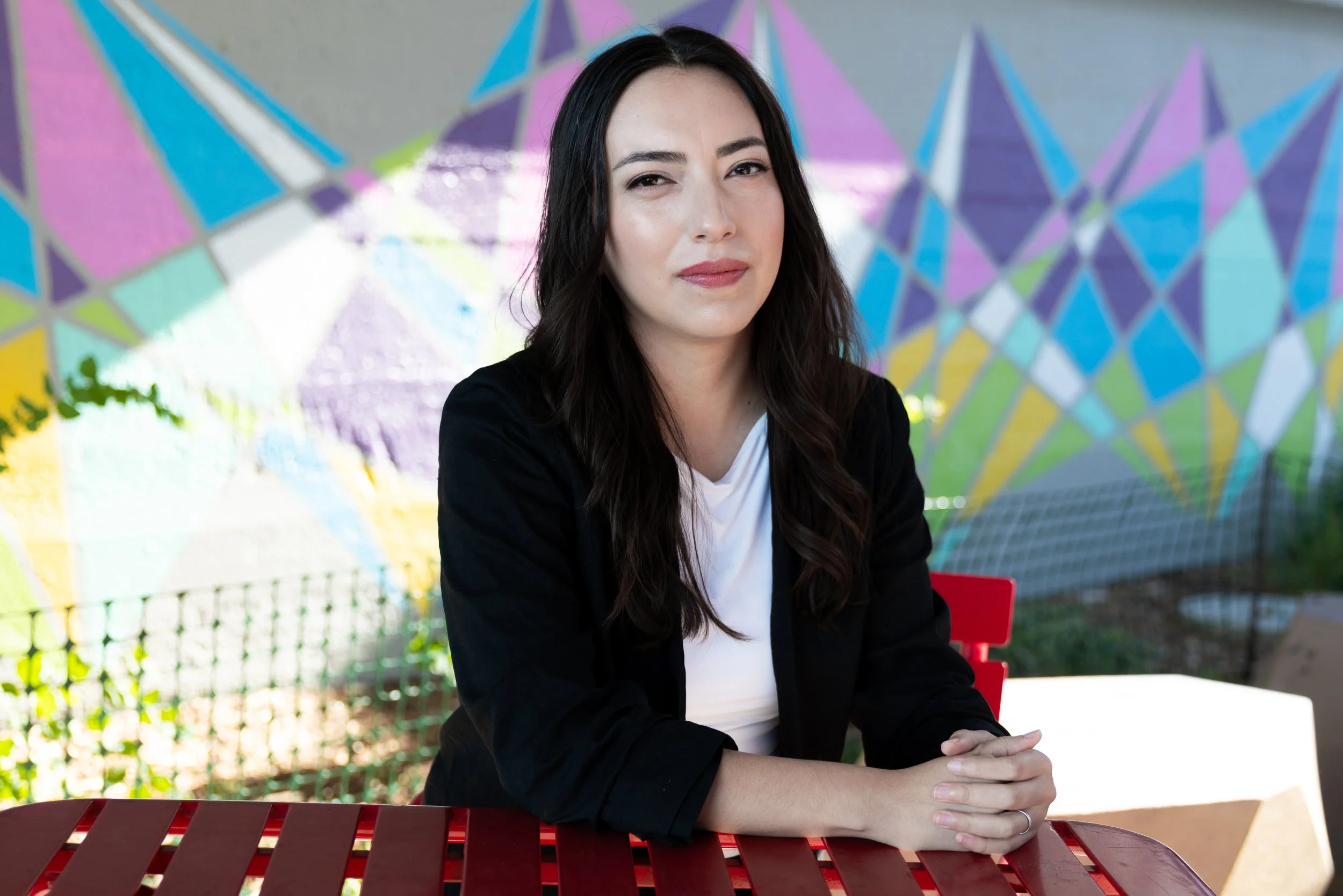A woman with long dark hair sitting at a red outdoor table with a colorful mural in the background.