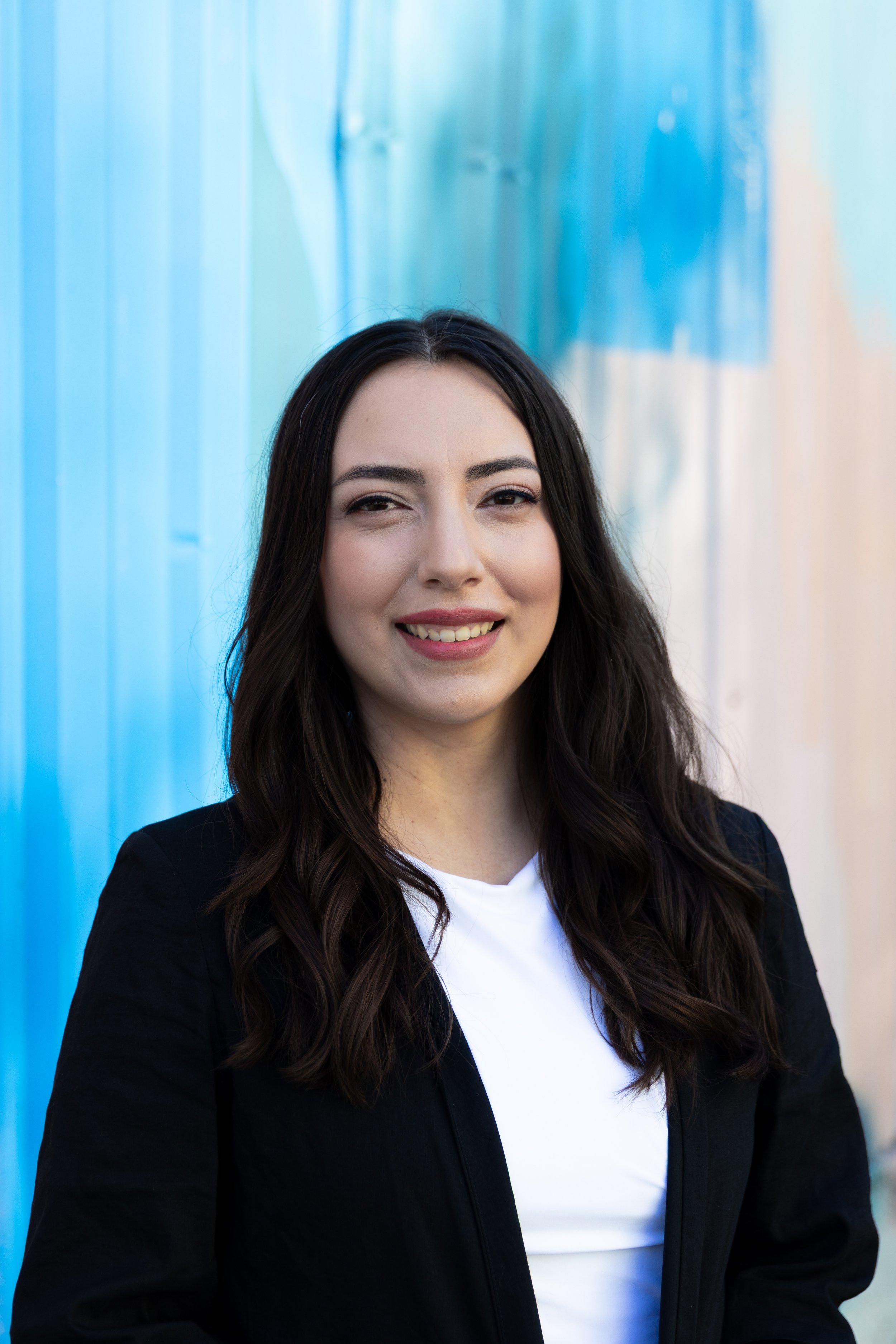 Portrait of a smiling woman with long dark hair, wearing a black jacket over a white shirt, standing against a colorful background.