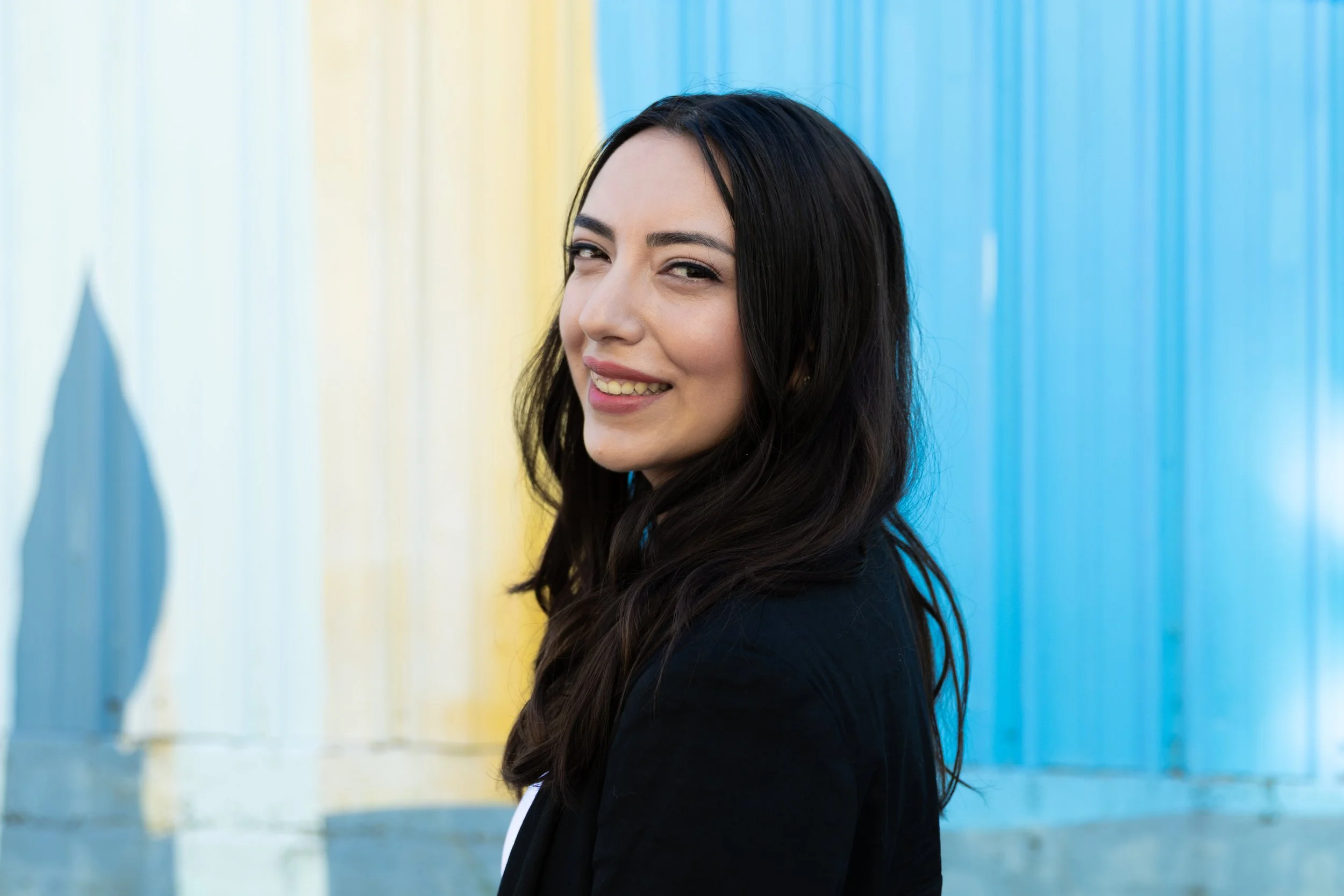 A young woman with long dark hair and a black blazer smiling at the camera, standing in front of a colorful background with vertical blue and yellow corrugated panels.