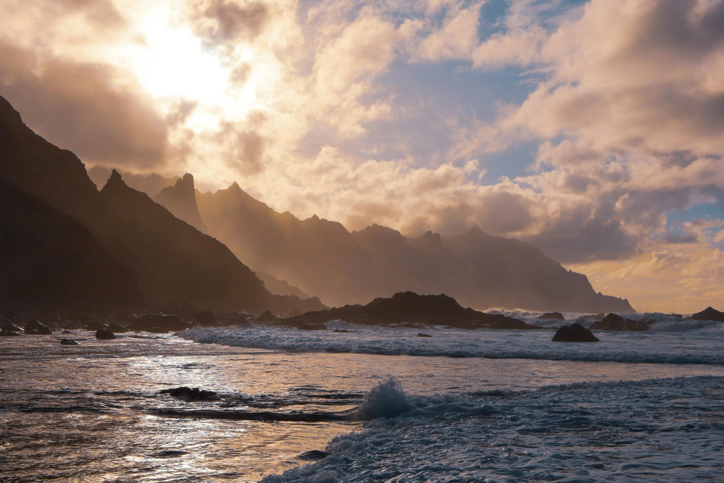 Sunset over rugged mountains and the ocean with waves crashing on the shoreline.
