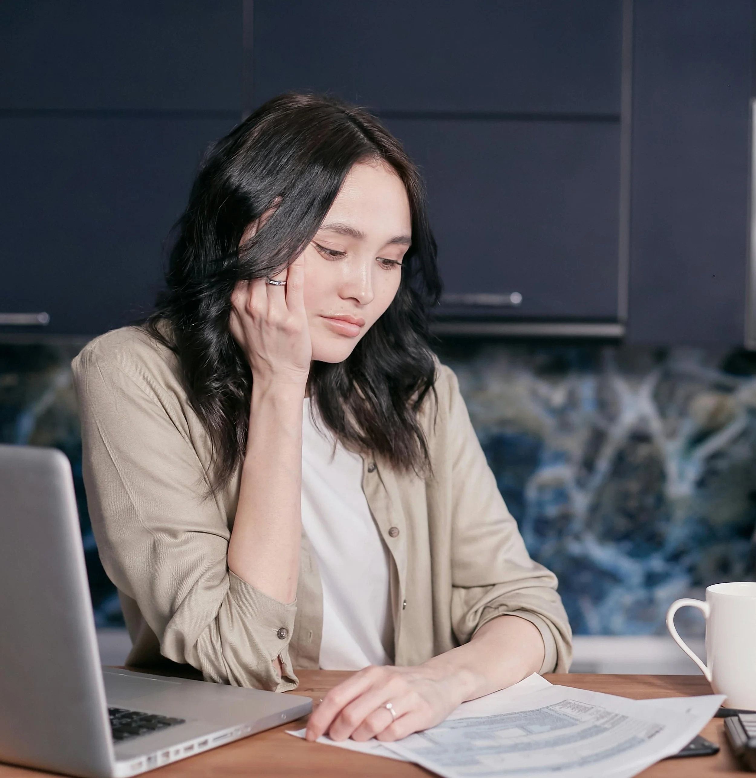 A woman sitting at a desk with a closed laptop, reading printed documents, with a coffee mug nearby, appearing thoughtful or concerned.