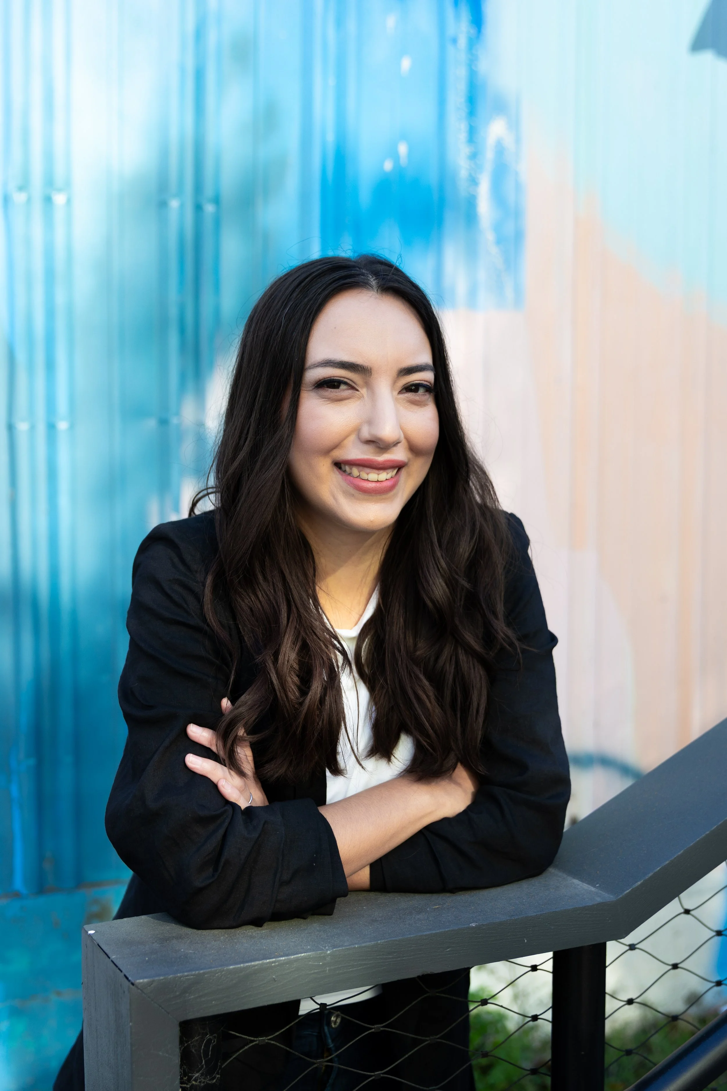 Young woman with long dark hair, wearing a black blazer, smiling while leaning on a grey fence, with a colorful, abstract blue background.