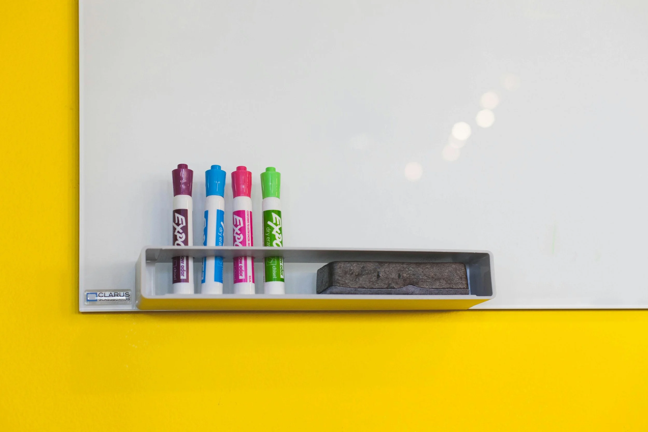 Whiteboard with four colorful markers and a grey eraser in a tray against a yellow wall.