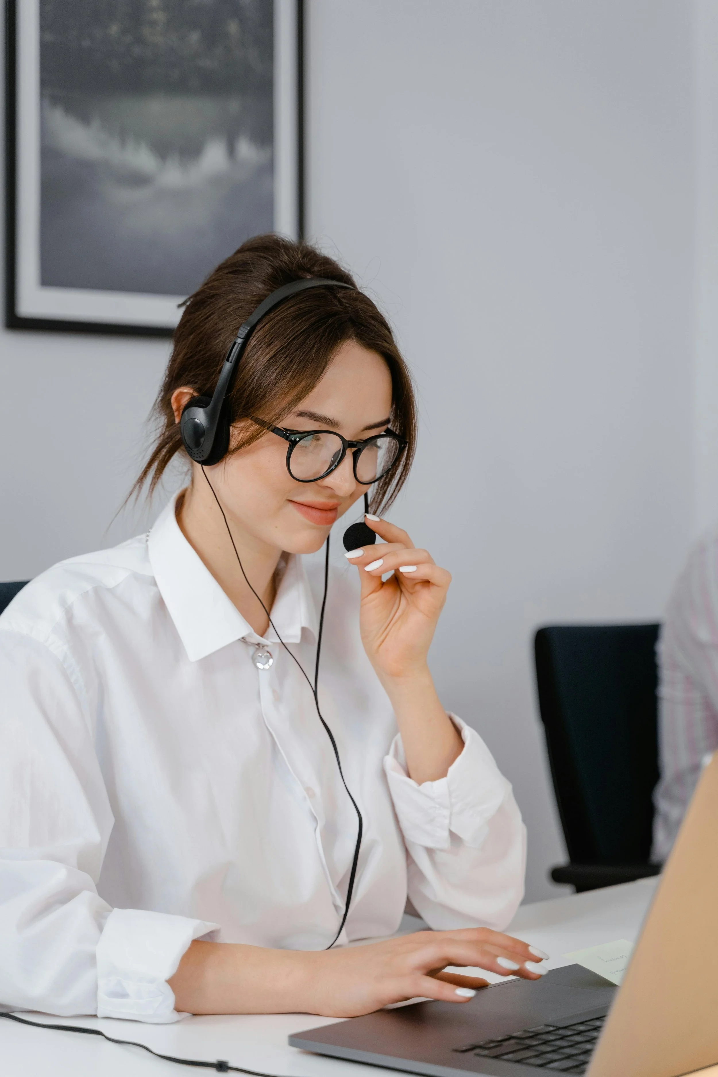 A woman with brown hair, glasses, and a white button-up shirt sitting at a table using a laptop. She is wearing a headset with a microphone, smiling, and looking at the laptop screen.