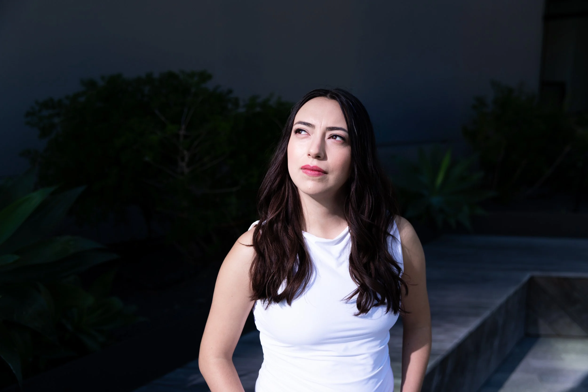 A young woman outdoors at night wearing a white sleeveless top, gazing thoughtfully upward, with dark green plants in the background.