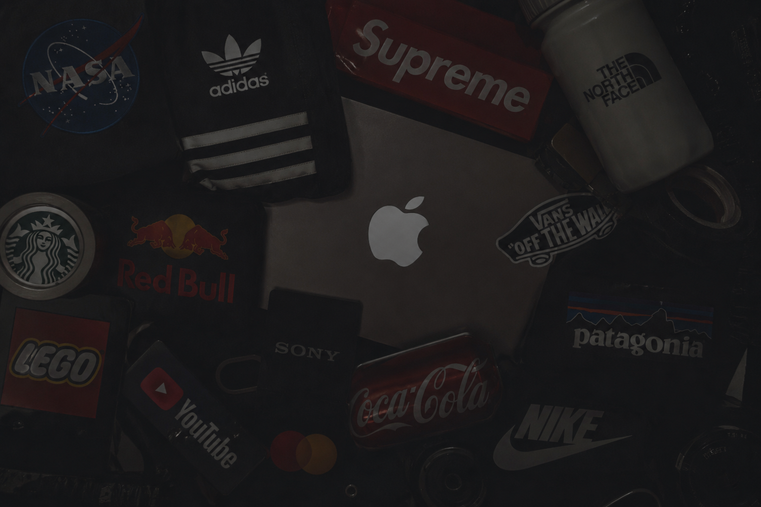 A silver MacBook surrounded by various branded items including a NASA patch, Adidas bag, Supreme box, The North Face bottle, Starbucks container, Red Bull logo, Vans 'Off The Wall' sticker, Patagonia patch, Nike logo, Coca-Cola tin, YouTube badge, and a watch.