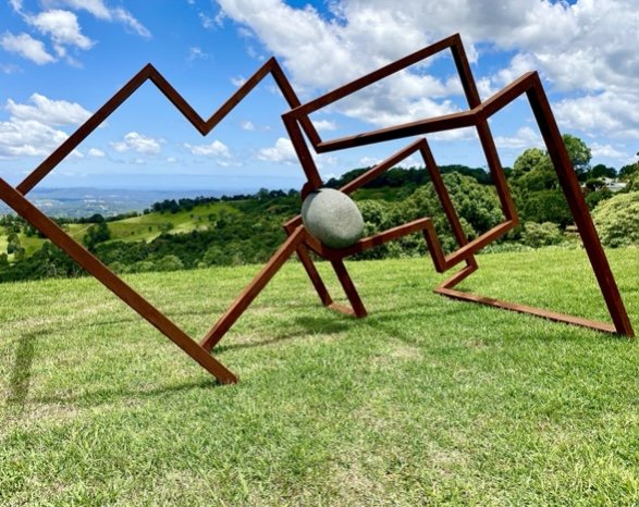 Abstract metal sculpture with geometric frames and a stone sphere on a grassy hilltop, under a blue sky with clouds.